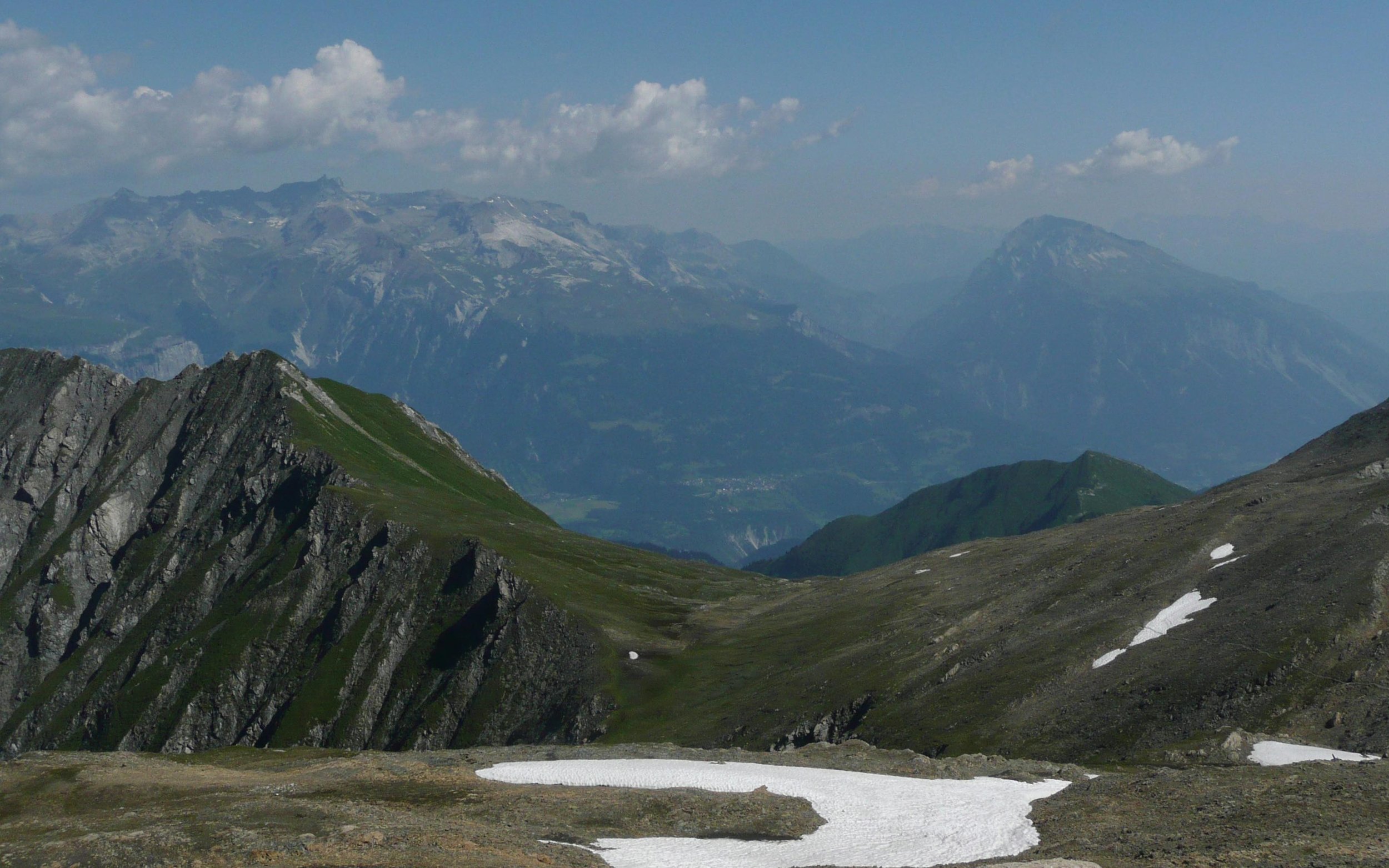 Berglandschaft mit grünen Hügeln, Felsen, kleinen Schneefeldern und Bergen im Hintergrund unter einem bewölkten Himmel.