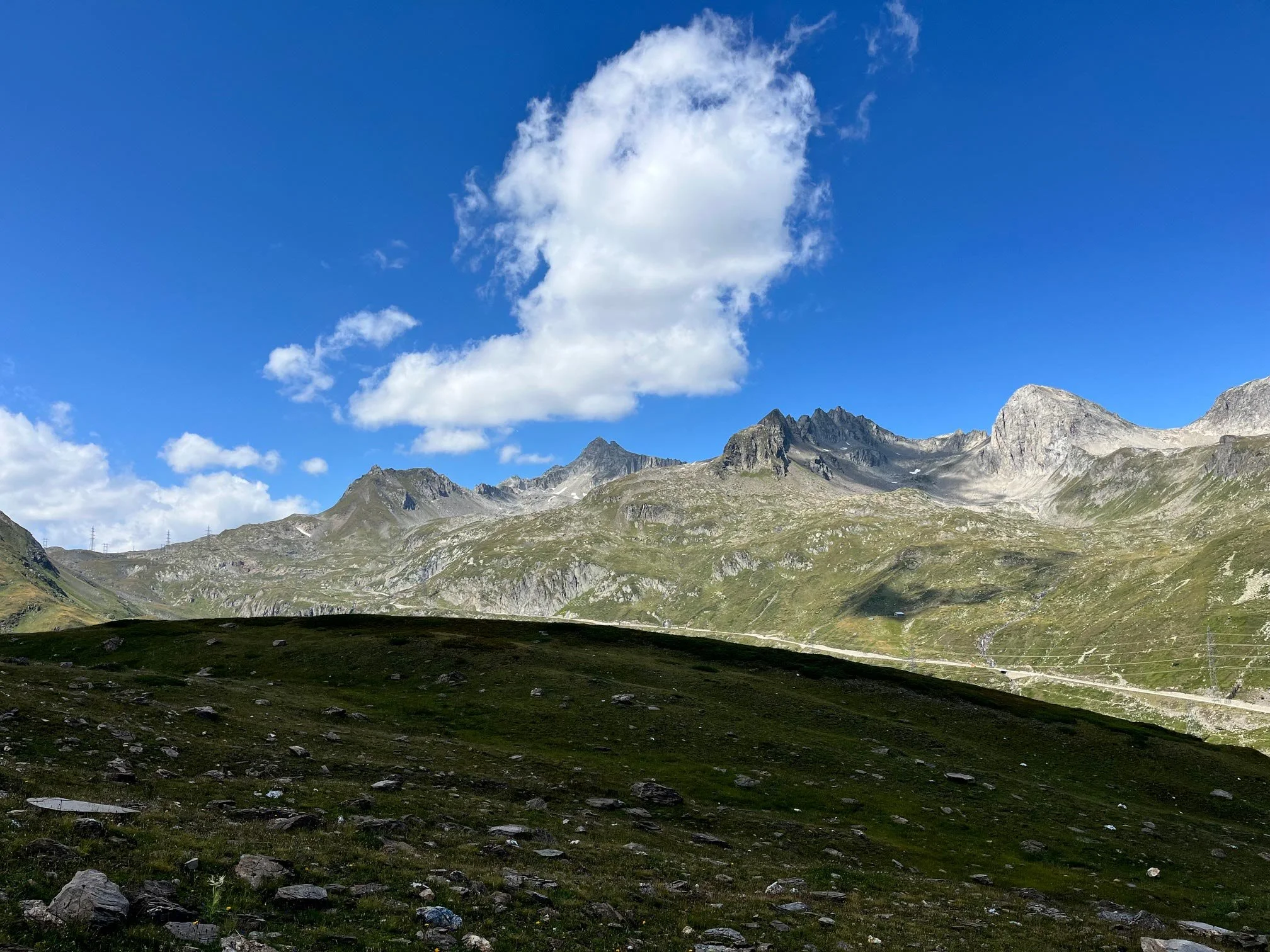 Berge in einer alpinen Landschaft unter einem blauen Himmel mit Wolken. Im Vordergrund liegt eine grüne Wiese mit Steinen.