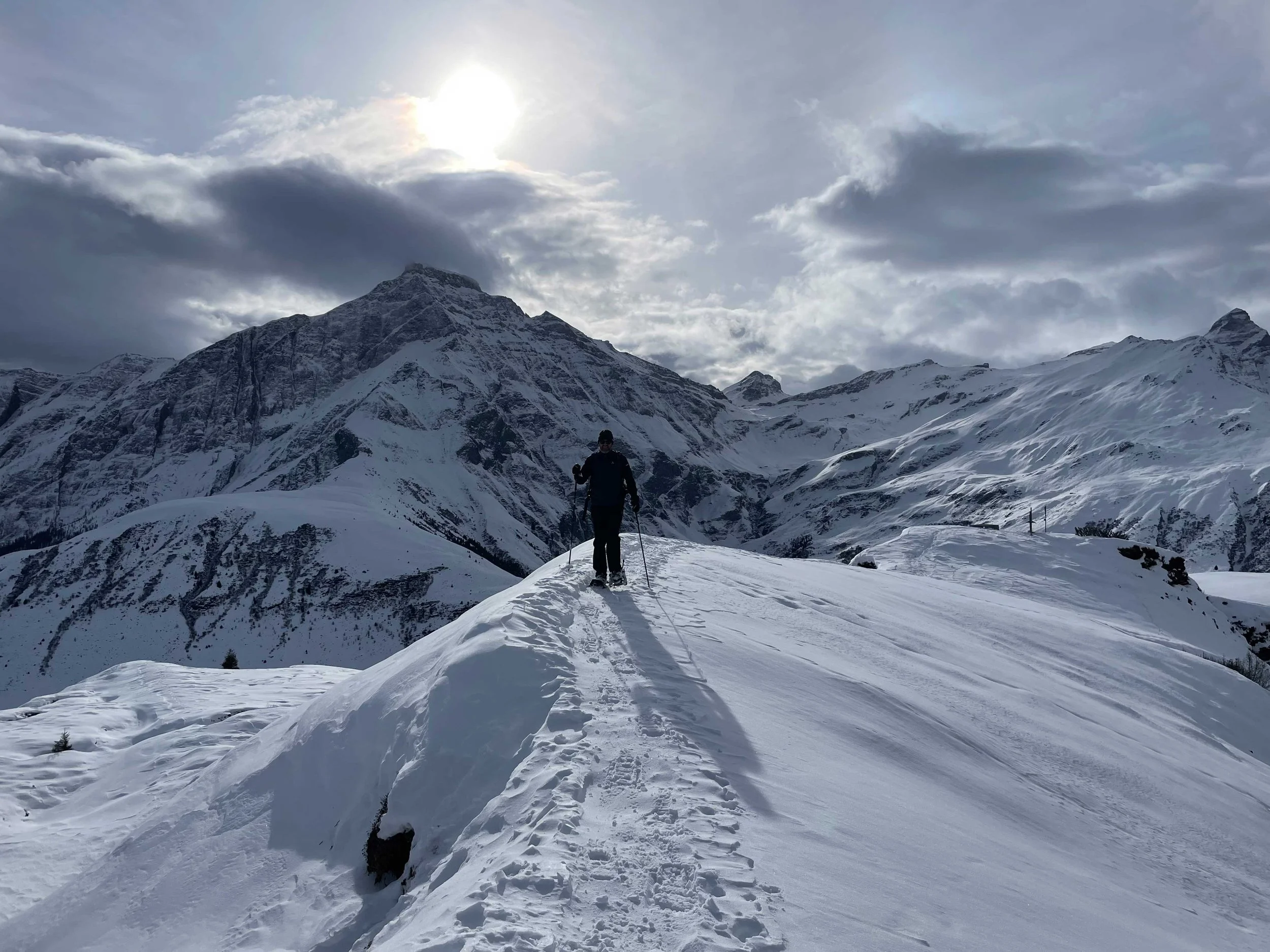 Eine Person wandert in verschneiter Berglandschaft bei bewölktem Himmel, mit Sonne im Hintergrund.