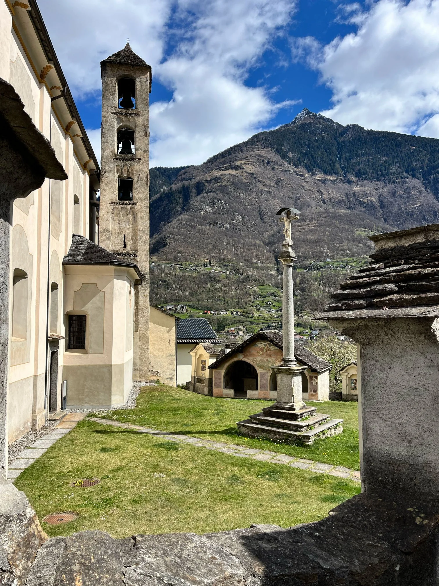 Historische Kirche mit Glockenturm, religiöse Statue auf Säule, kleine Kapellen im Grünen, Berge im Hintergrund, klarer Himmel mit Wolken.