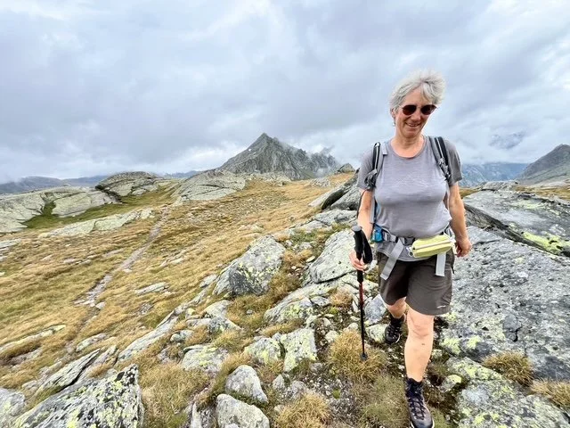 Eine Frau wandert auf einem Bergpfad in einer alpinen Landschaft, umgeben von Steinen und Gräsern, mit Bergen im Hintergrund, trägt Sonnenbrille, Wanderausrüstung und einen Wanderstock.