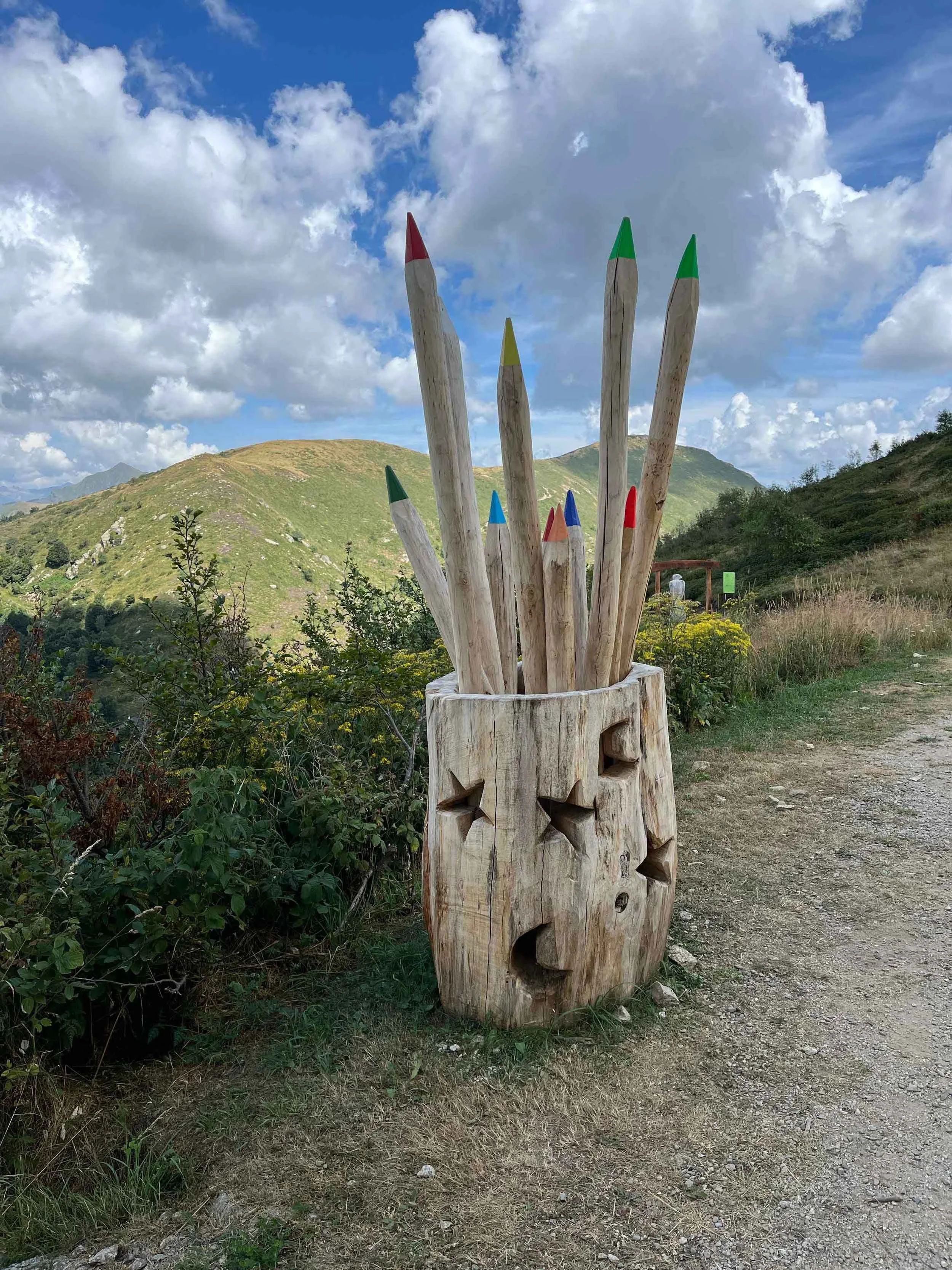 Ein großes Holzskulptur, bestehend aus mehreren senkrecht stehenden Holzbalken mit farbigen Spitzen, in einer natürlichen Landschaft mit grünen Hügeln und einem bewölkten Himmel im Hintergrund.