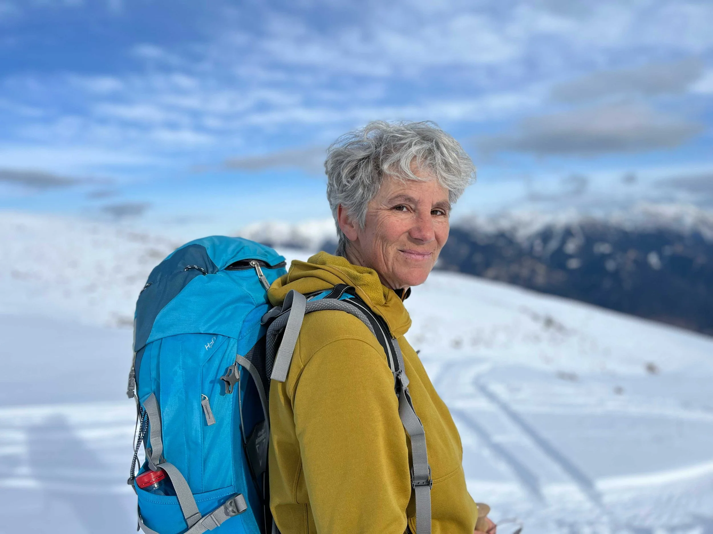 Lächelnde Frau mit grauem Haar trägt eine gelbe Jacke und einen großen blauen Rucksack in verschneiter Berglandschaft mit Wolken am Himmel.