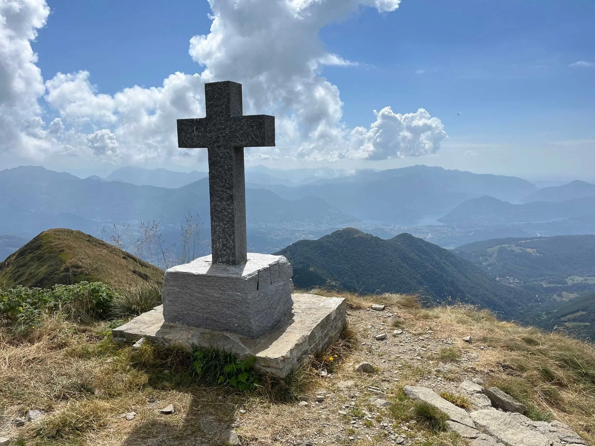 Stein Kreuz auf einem Berggipfel mit Blick auf eine Bergkette und Wolken am Himmel.