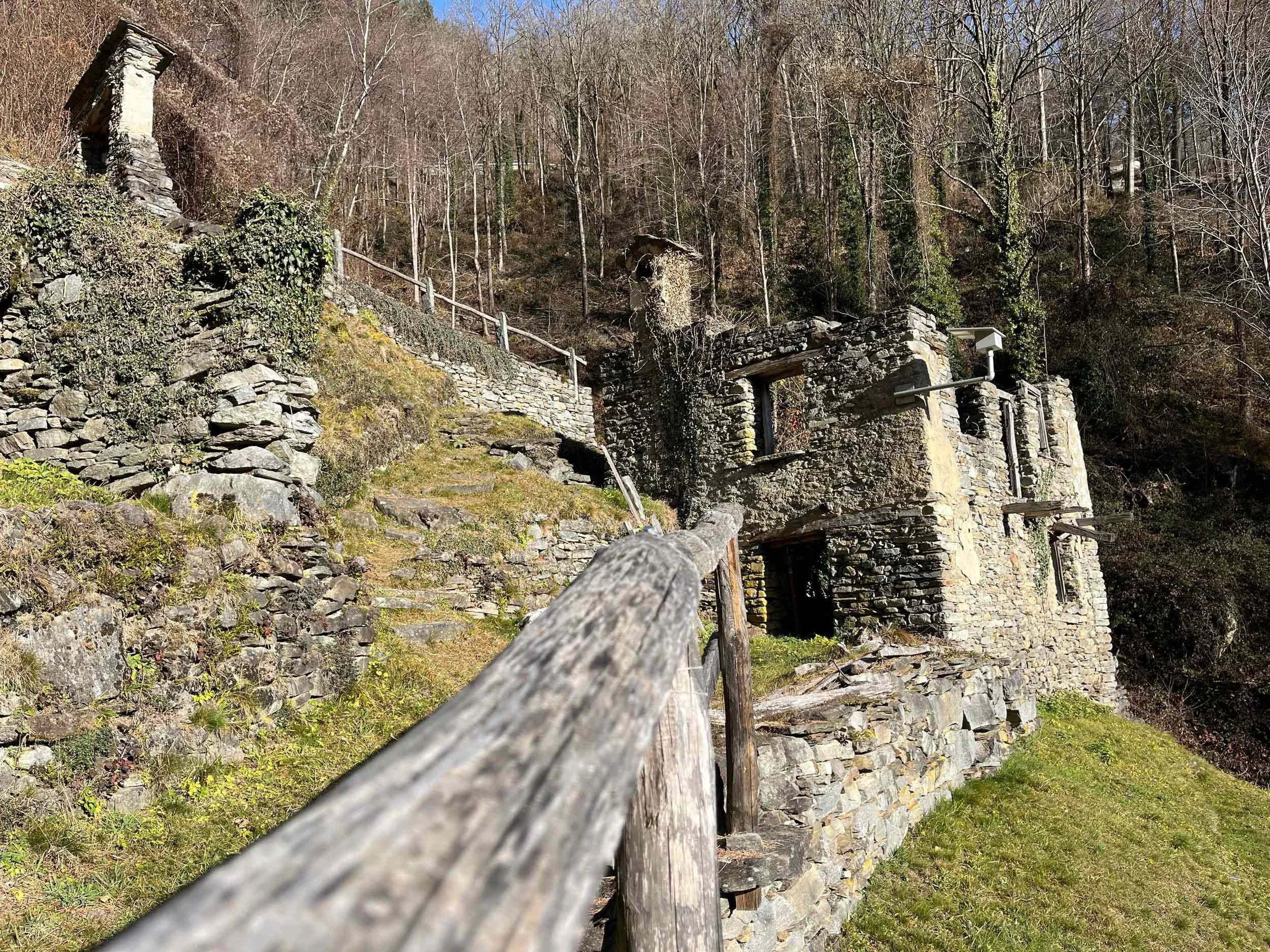 Ein steinerner Weg führt an einem alten Baumhaus im Wald entlang, im Hintergrund sind Bäume ohne Blätter zu sehen.