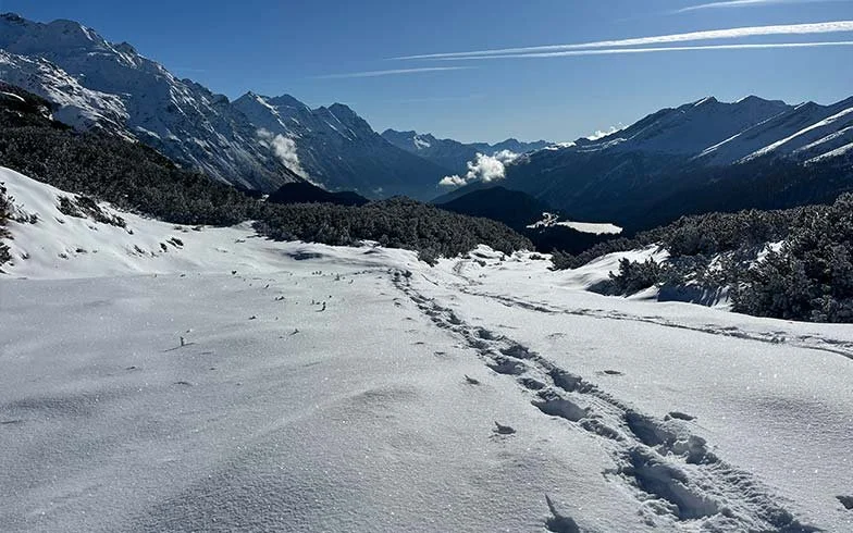 Verschneite Berge und Tal mit Fußspuren im Schnee, blauer Himmel mit Wolken und Kondensstreifen.