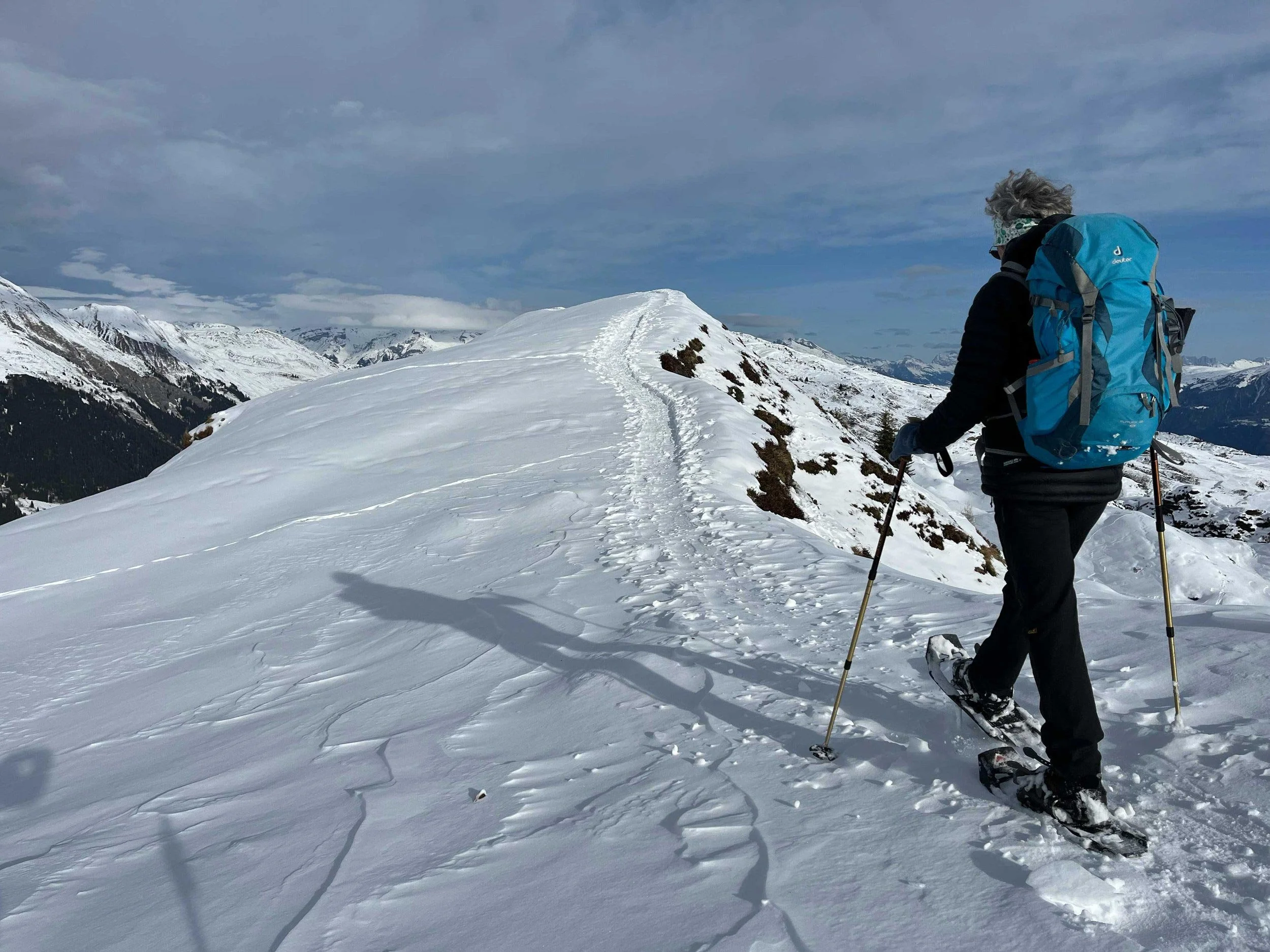 Eine Person mit Winterkleidung, Skischuhen und einem blauen Rucksack beim Skifahren auf verschneitem Bergkamm mit Blick auf schneebedeckte Berge und Wolken am Himmel.