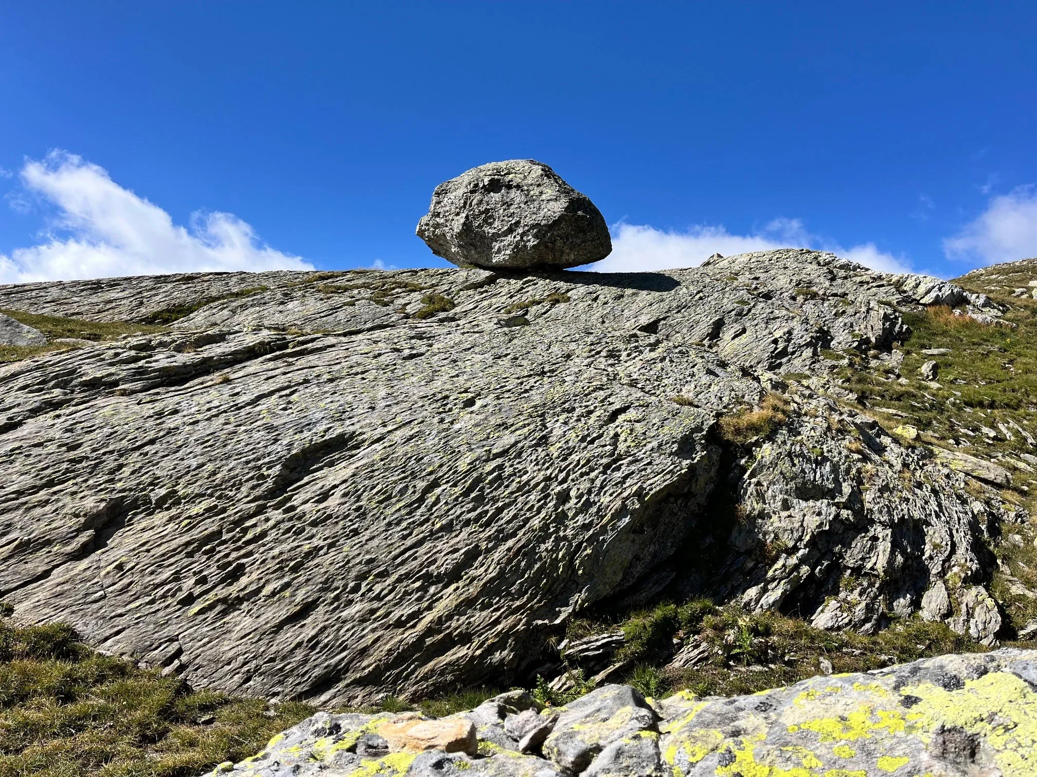 Ein einzelner großer Felsen auf einer felsigen Berghochebene, mit blauer Himmel und einigen Wolken im Hintergrund. Gras und kleinere Steine im Vordergrund.