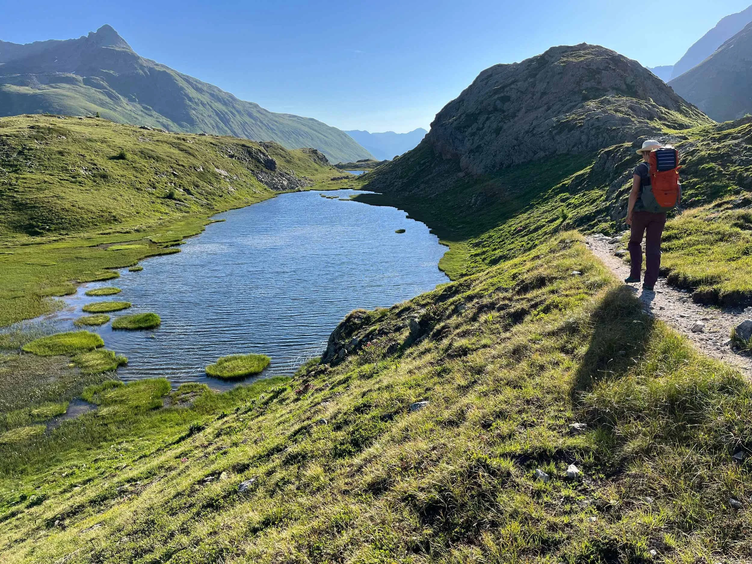Ein Wanderer mit Rucksack unterwegs entlang eines Flusses in einer bergigen Landschaft mit grünen Hügeln und hohen Bergen im Hintergrund bei sonnigem Wetter.