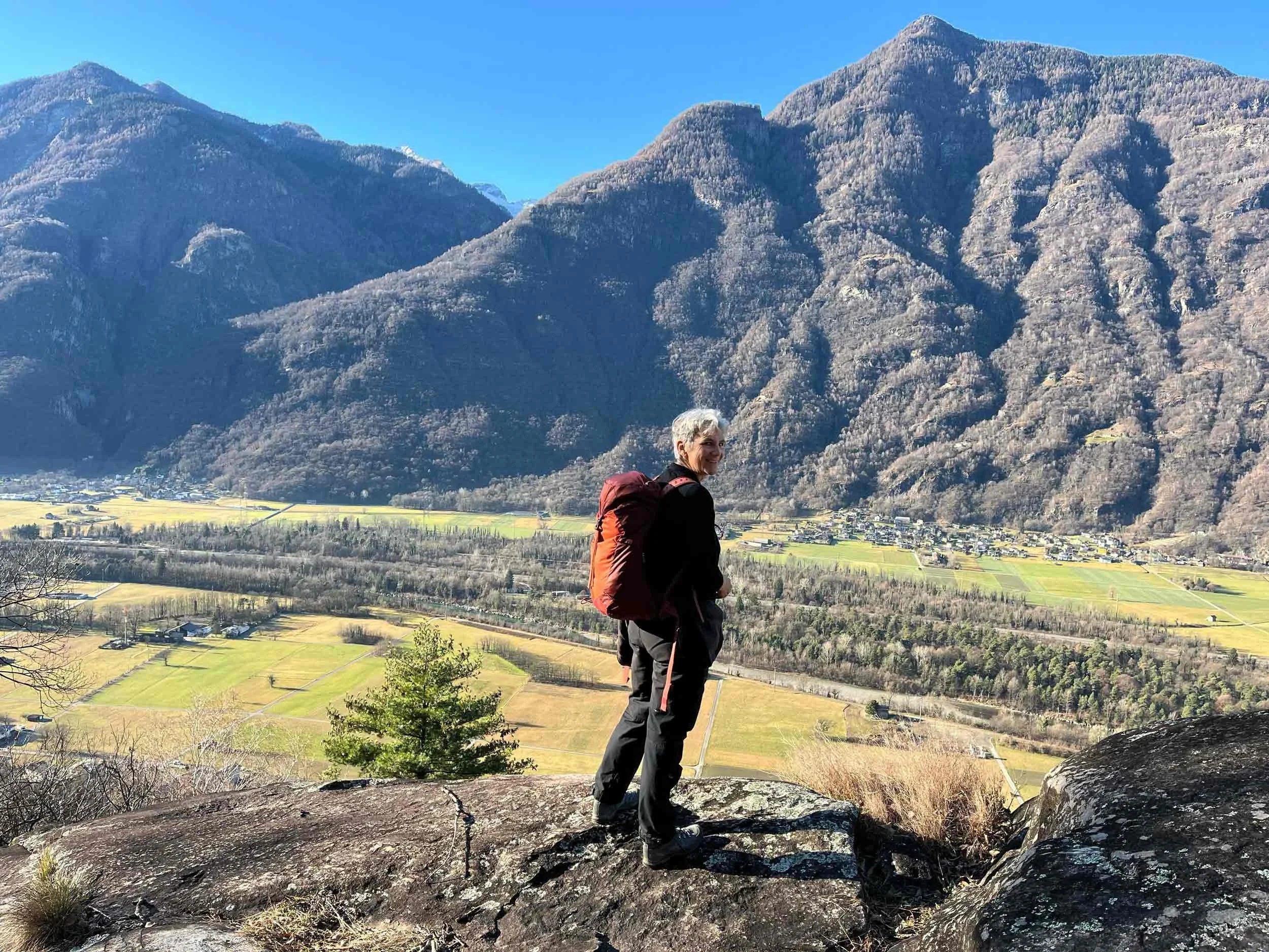 Eine Frau mit grauen Haaren wandert in den Bergen. Im Hintergrund sind hohe, schneebedeckte Berge und eine fruchtbare, grüne Ebene mit einem kleinen Dorf.