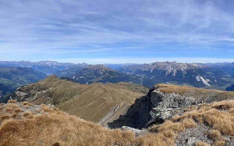 Berglandschaft mit Gras, Felsen und entfernten Gipfeln unter einem blauen Himmel.