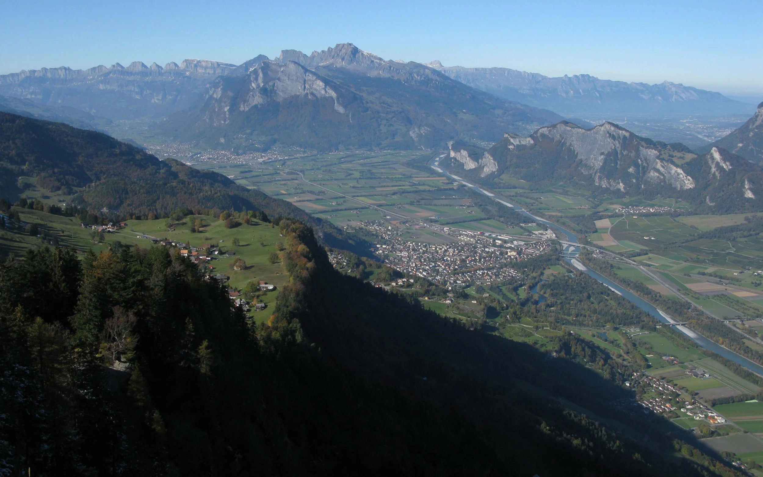 Berglandschaft mit hohen Bergen, Tälern, Fluss und Siedlungen im Tal, im Vordergrund Wälder.