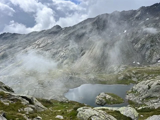 Ein Bergsee inmitten von felsigem, alpinem Gelände, umgeben von steilen Hügeln und teilweise bewölktem Himmel.