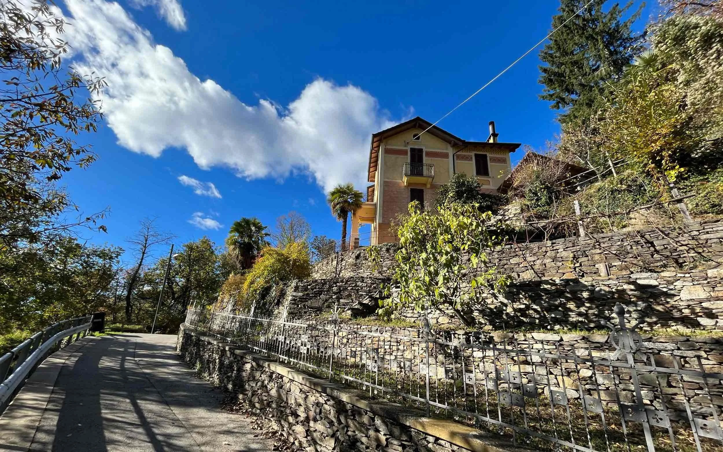 Ein Haus auf einem Hügel, umgeben von Bäumen, mit einer steinernen Mauer und einem Weg, im blauen Himmel mit einigen Wolken.