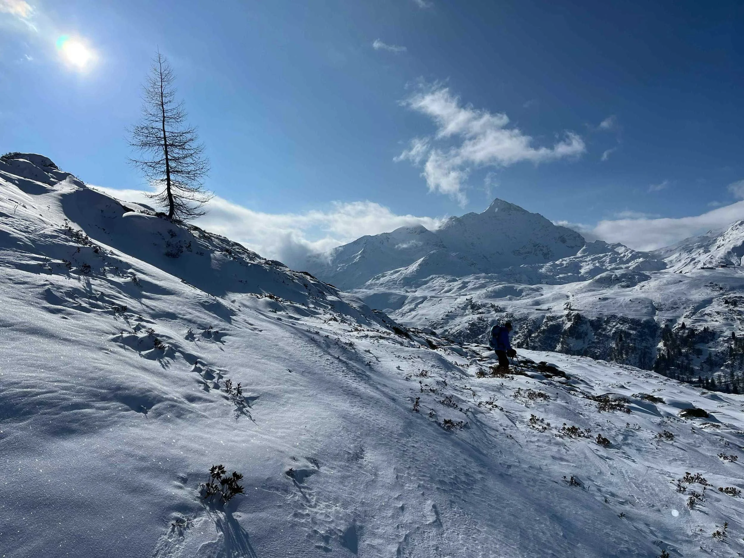 Schneebedeckte Berge und eine einsame, kahle Baumlücke mit einem kleinen Schneemann, umgeben von klarem blauen Himmel und einigen Wolken.