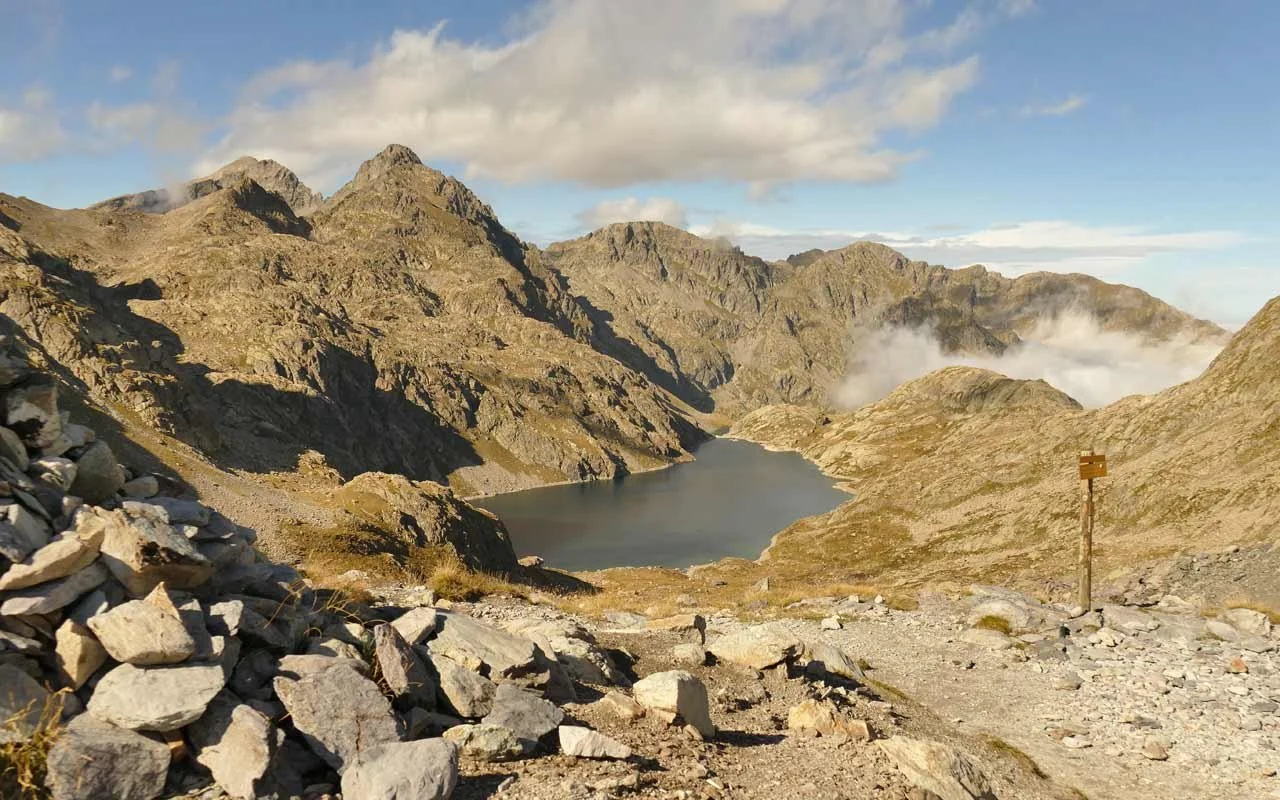 Bergsee in einer felsigen Gebirgslandschaft mit Bergen im Hintergrund und einer Wegmarkierung im Vordergrund.