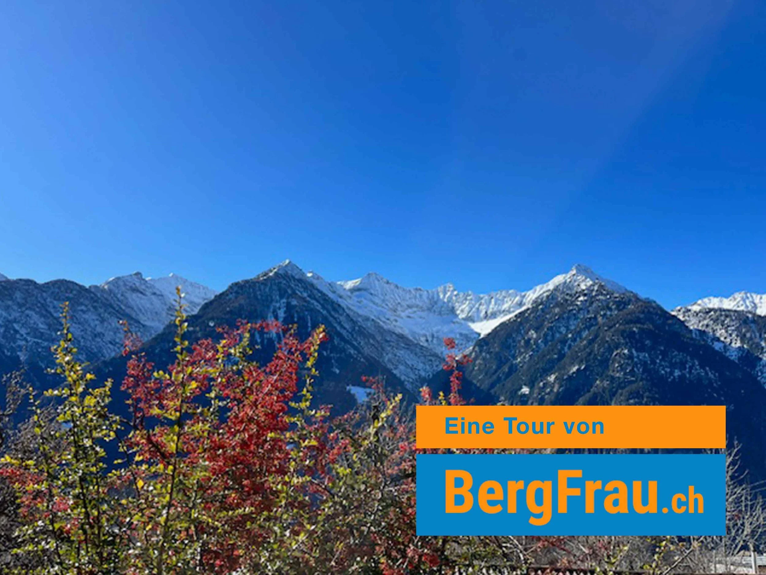 Berglandschaft mit verschneiten Gipfeln, blauer Himmel und bunte Bäume im Vordergrund, Textüberlagerung mit 'Eine Tour von BergFrau.ch'.
