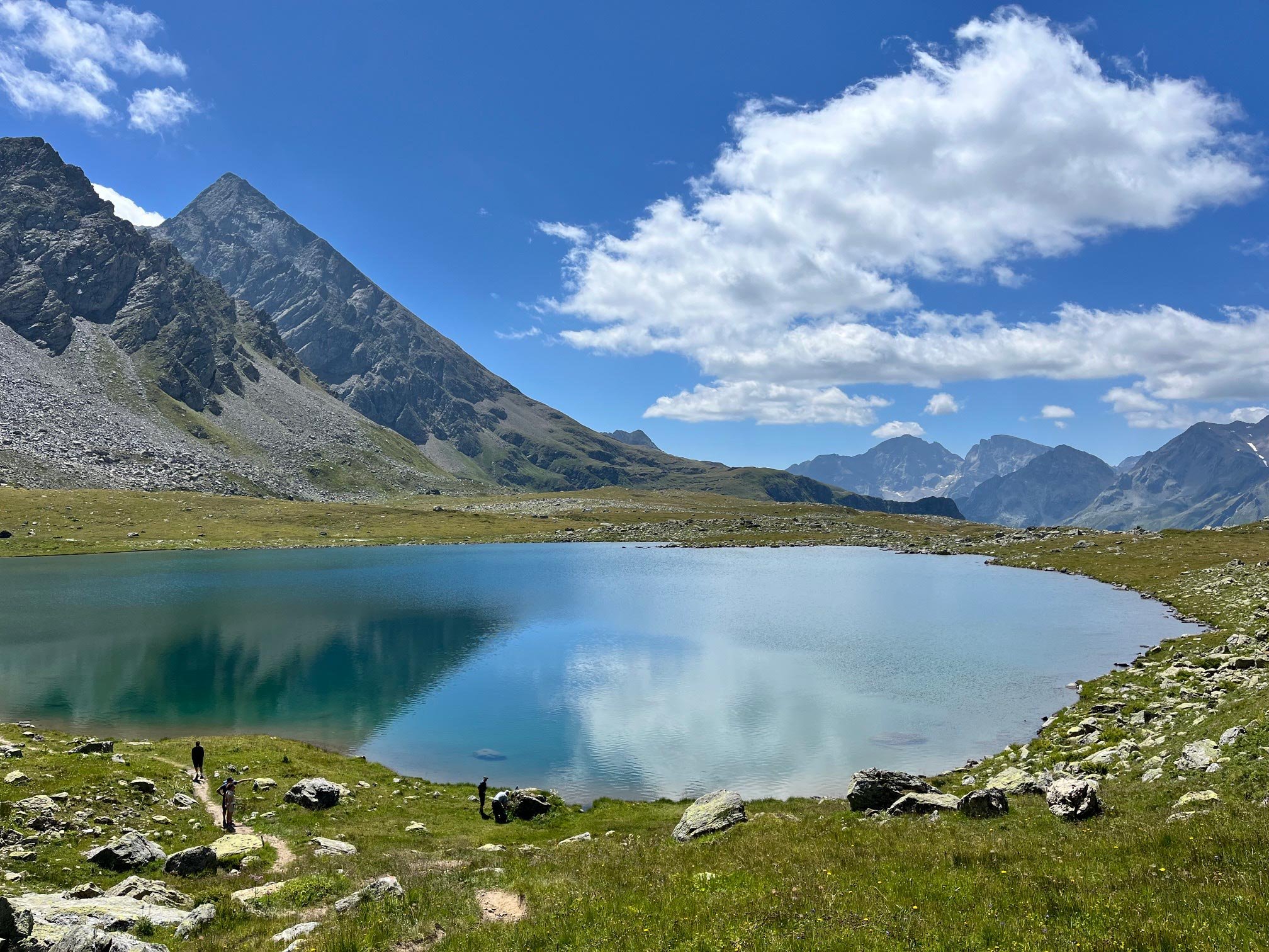 Bergsee mit klarer Wasseroberfläche, umgeben von grünem Gras und Felsen, vor hohen Bergen unter einem blauen Himmel mit Wolken, mit einigen Menschen am Ufer und auf einem Weg.