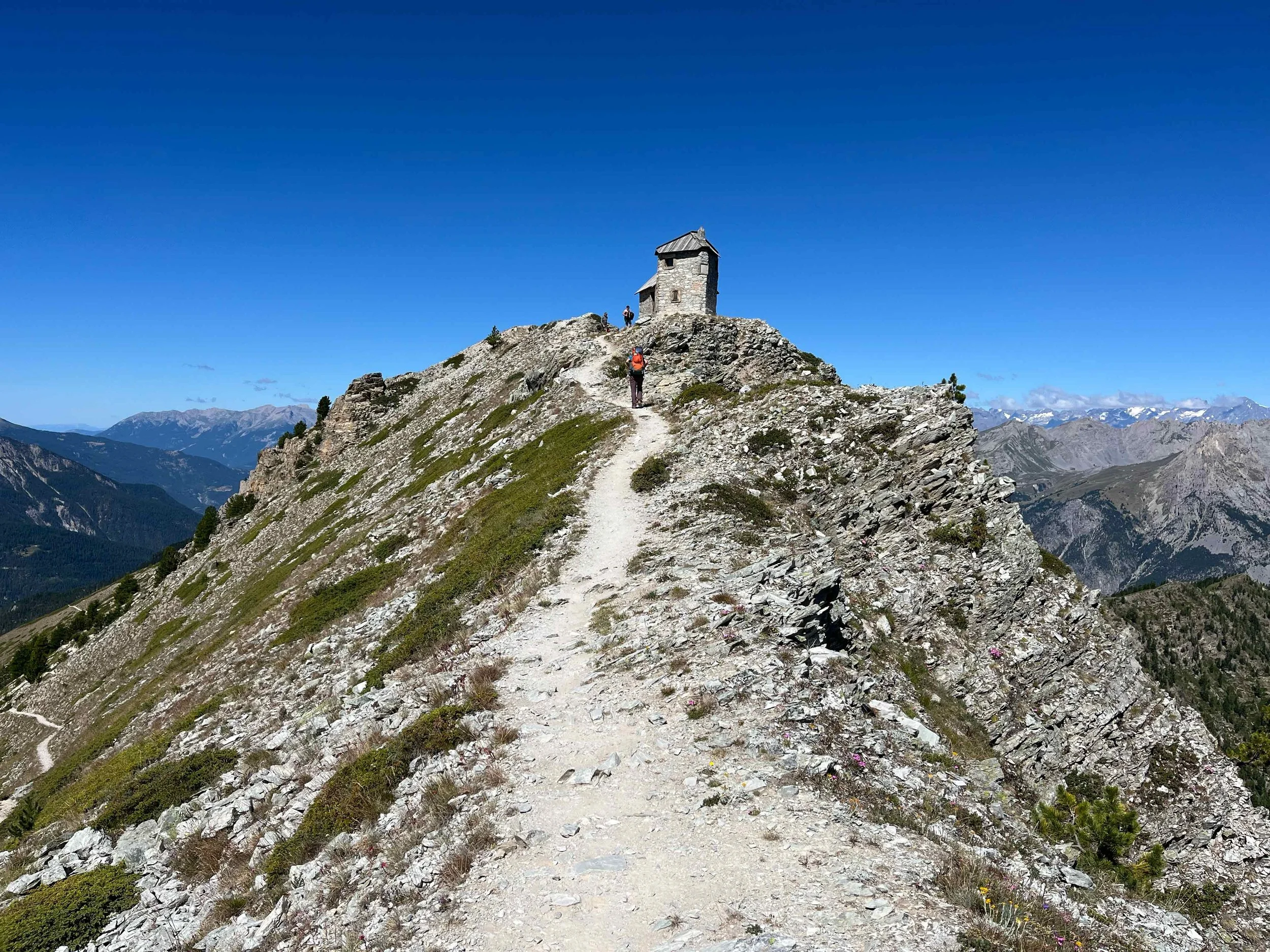 Wanderer wandert auf einem schmalen Bergpfad mit Blick auf eine kleine Steinhütte auf dem Gipfel und bergige Landschaft im Hintergrund.