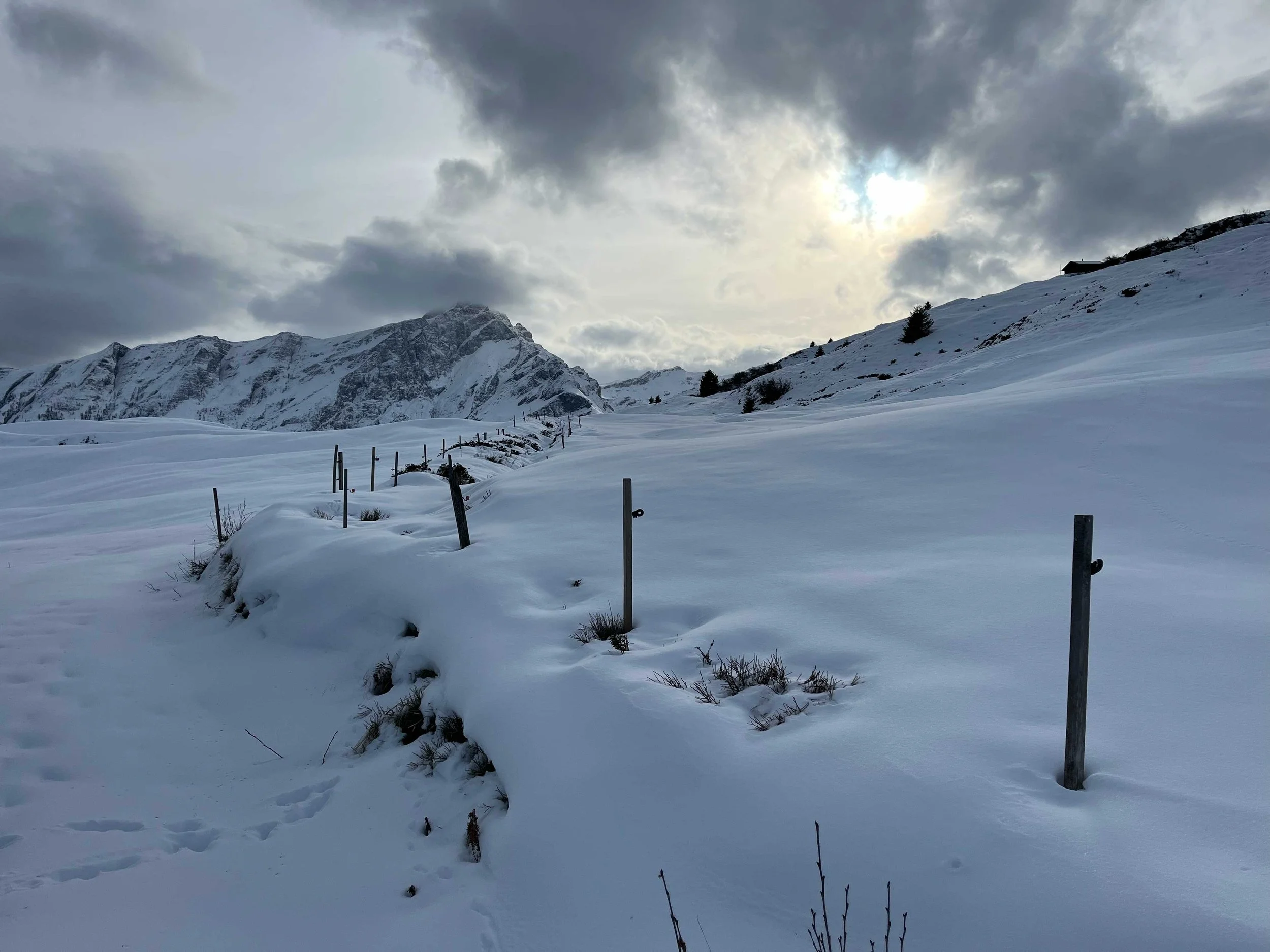 Schneebedeckte Berge und ein Hügel, Wolken am Himmel, eine dünne Schneeschicht auf dem Boden, einige verstreute Büsche und Stangen in der Schneedecke, im Hintergrund sind Berge und der Himmel mit teilweise sonnigen Wolken.