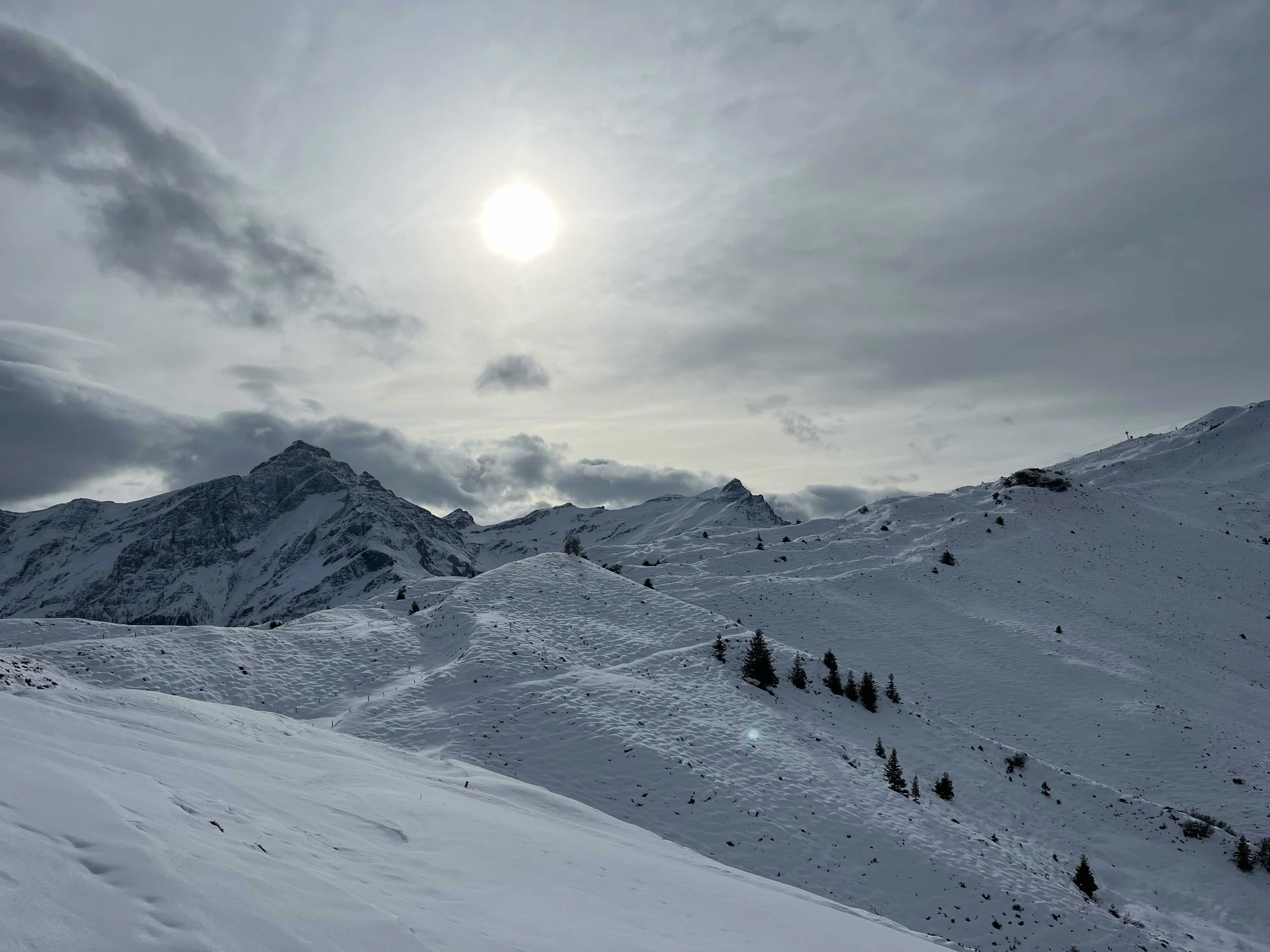 Schneebedeute Berge unter bewölktem Himmel mit der Sonne sichtbar.