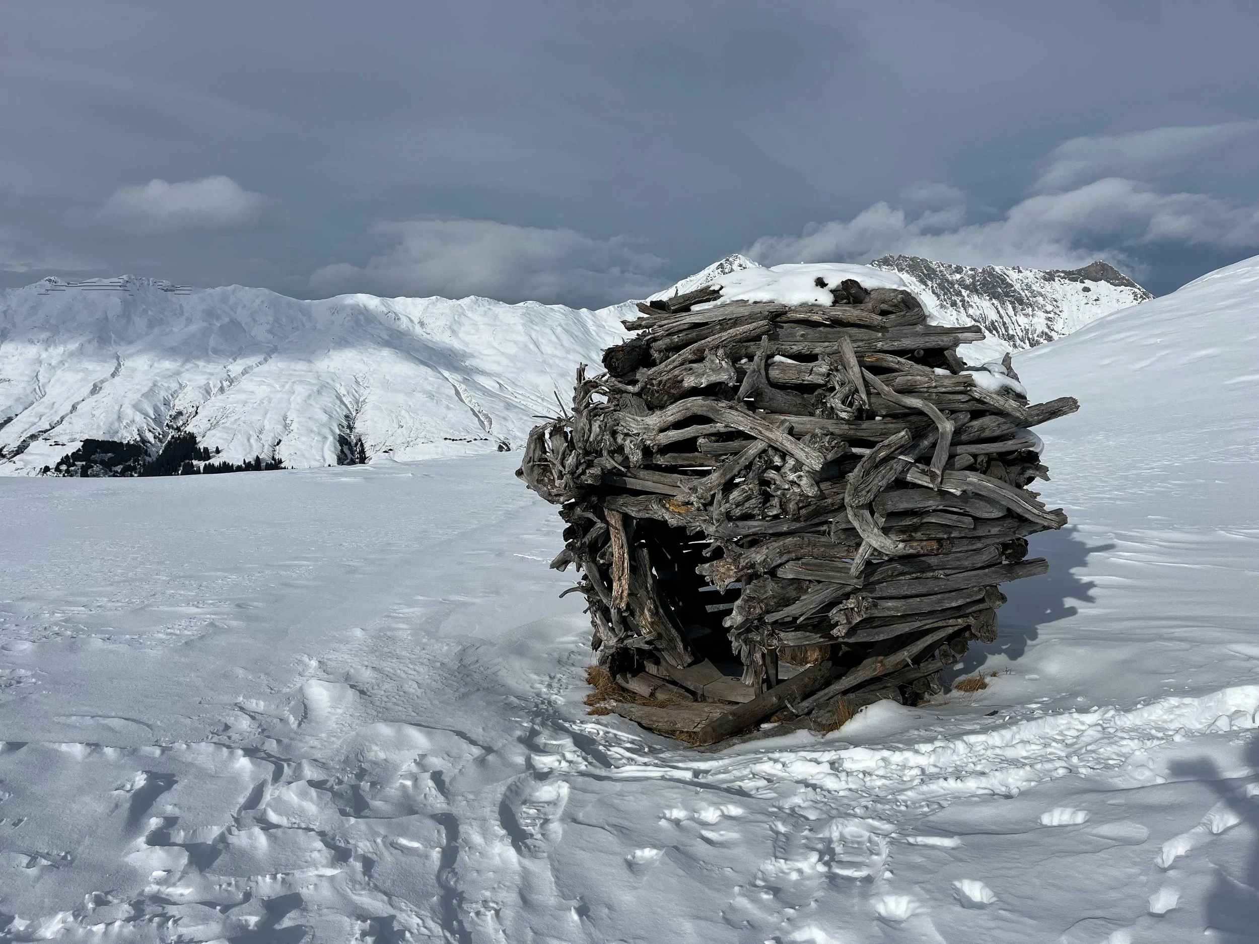 Ein kleines Holzhaus aus zusammengesetzten Holzstücken in einer verschneiten Schneelandschaft mit Bergen im Hintergrund.