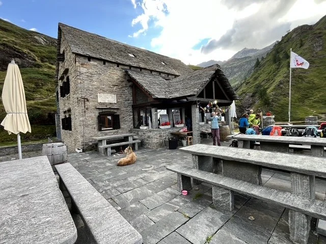 Berghütte Capanna Basodino mit Steinterrasse, Berglandschaft im Hintergrund und Menschen, die draußen sitzen und sich unterhalten.