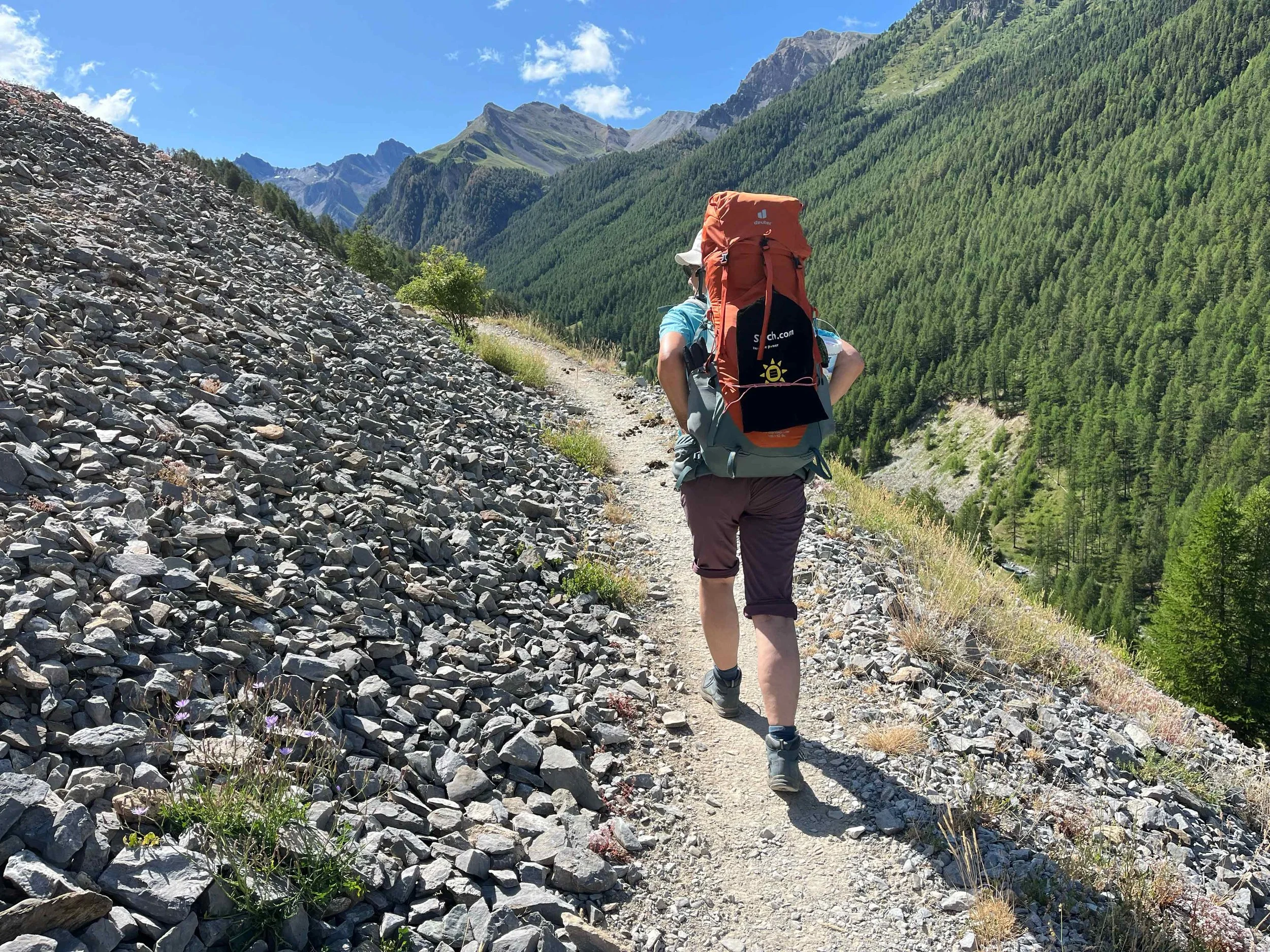 Eine Wanderin mit Rucksack auf einem schmalen Bergpfad im Gebirge, umgeben von grünen Wäldern und hohen Bergen unter blauem Himmel.
