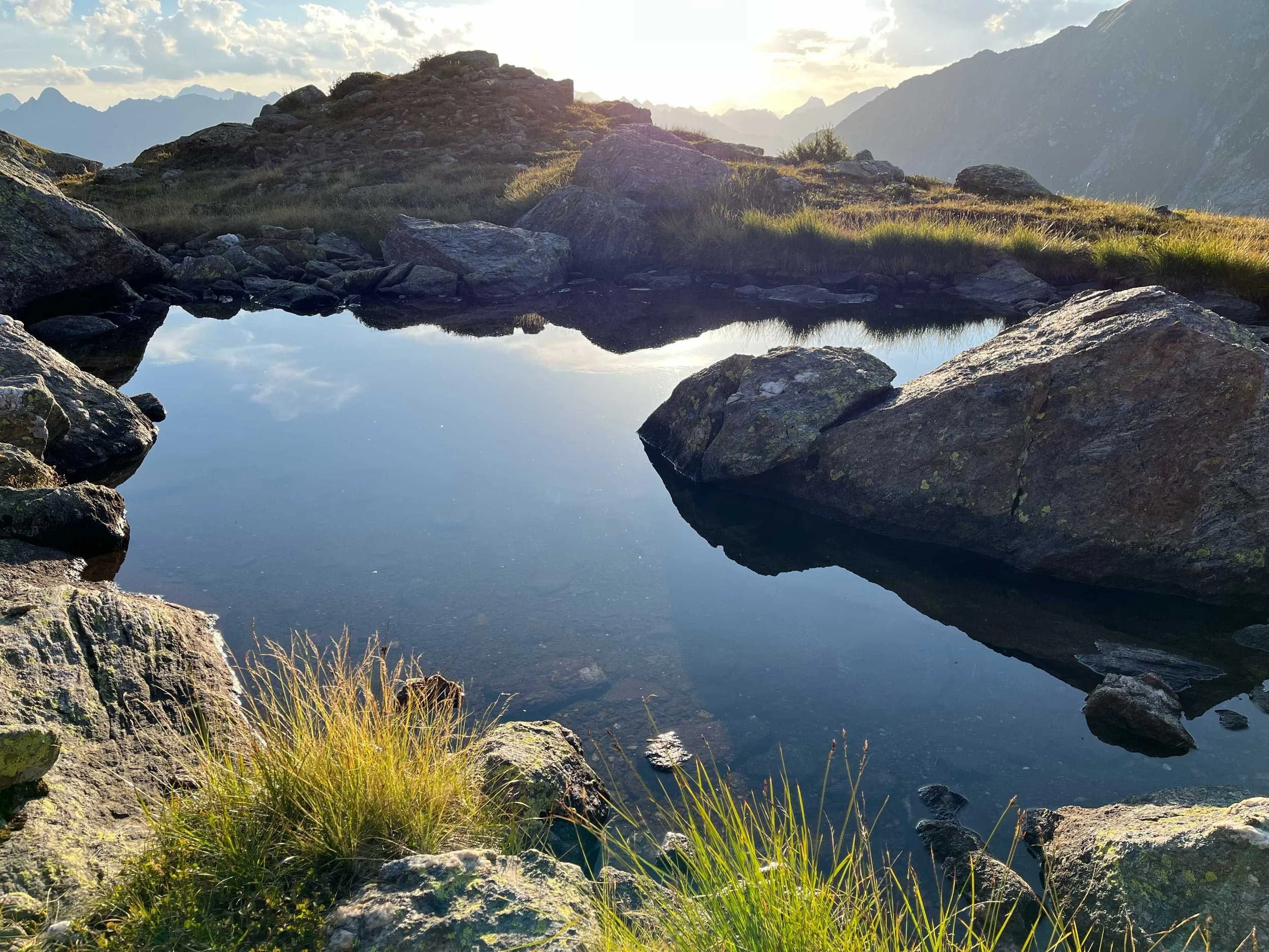 Ein See in den Bergen, umgeben von Felsen und Gras, mit Himmel und Wolken im Hintergrund, bei Sonnenuntergang oder Sonnenaufgang.