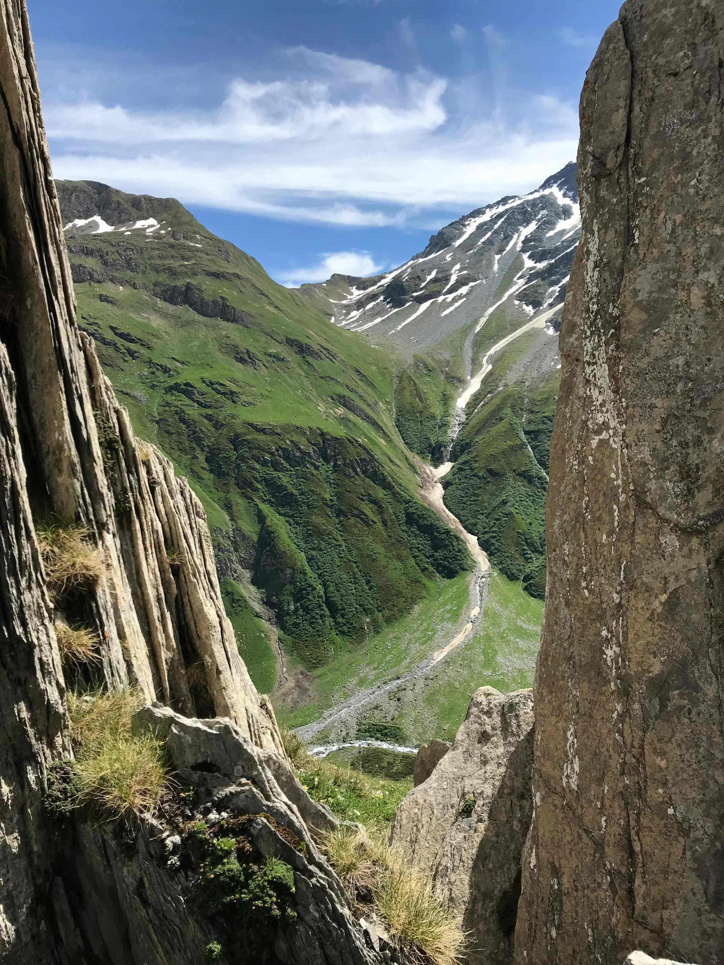 Berglandschaft mit grünen Hügeln, Schneeresten auf den Gipfeln, Wasserläufen und Wolken am Himmel, sichtbar durch eine Felsspalte.