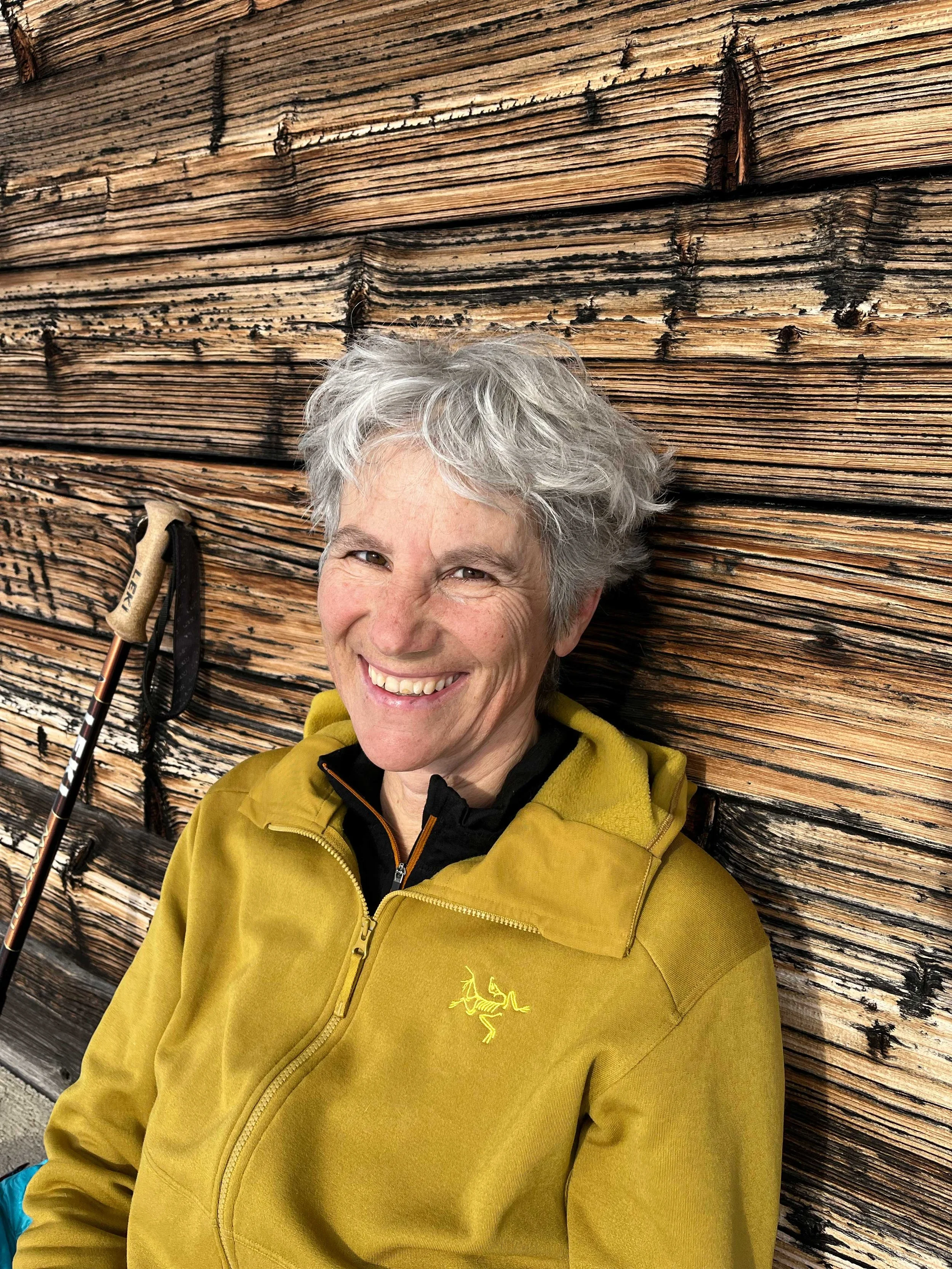 Ein lächelnder älterer Mann mit grauem, lockigem Haar, der in einem gelben Jäckchen vor einer rustikalen Holzwand sitzt, neben einem Wanderstock.