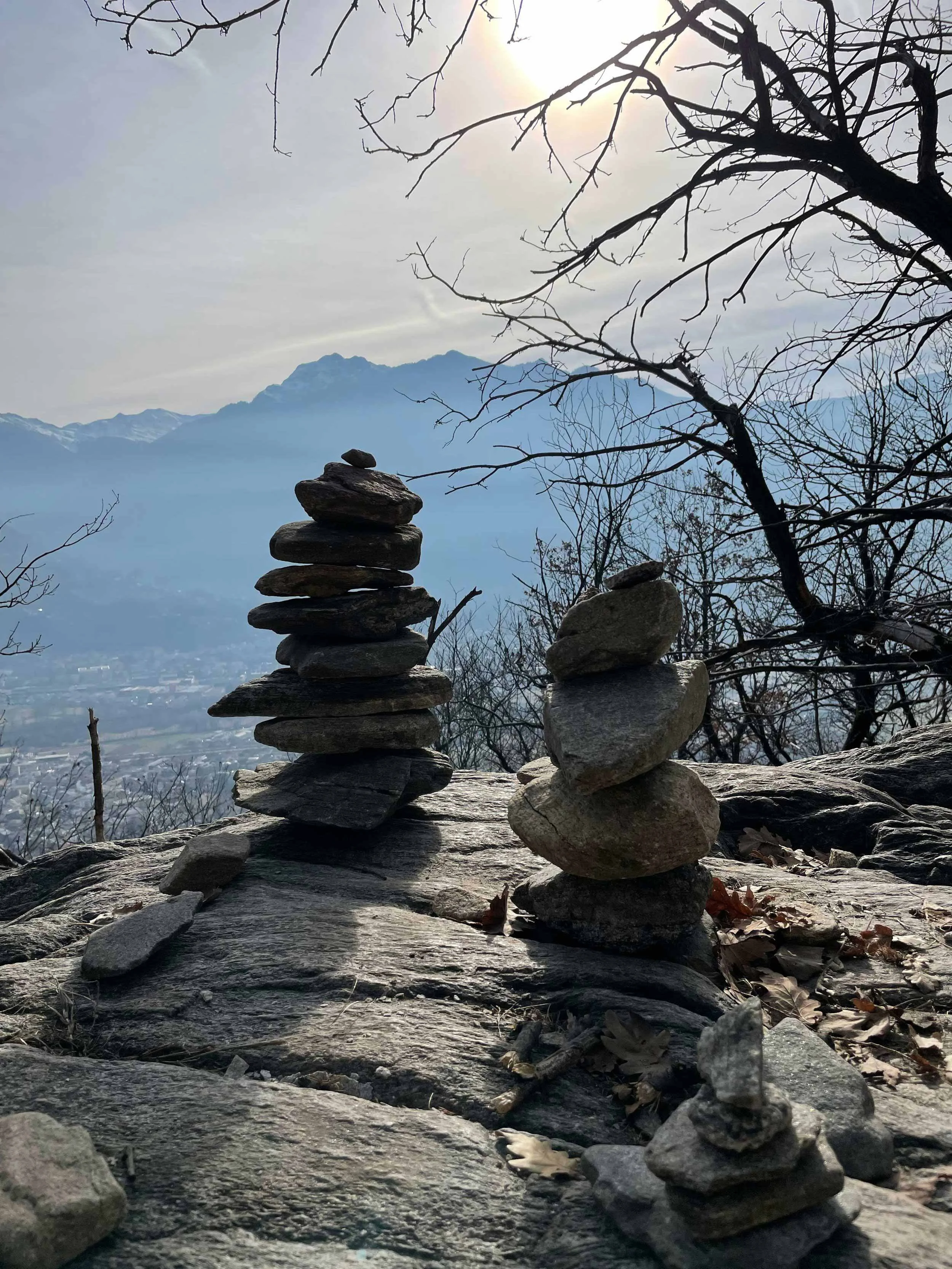Steinmännchen im Vordergrund, Berge im Hintergrund, kahle Bäume und bewölkter Himmel.