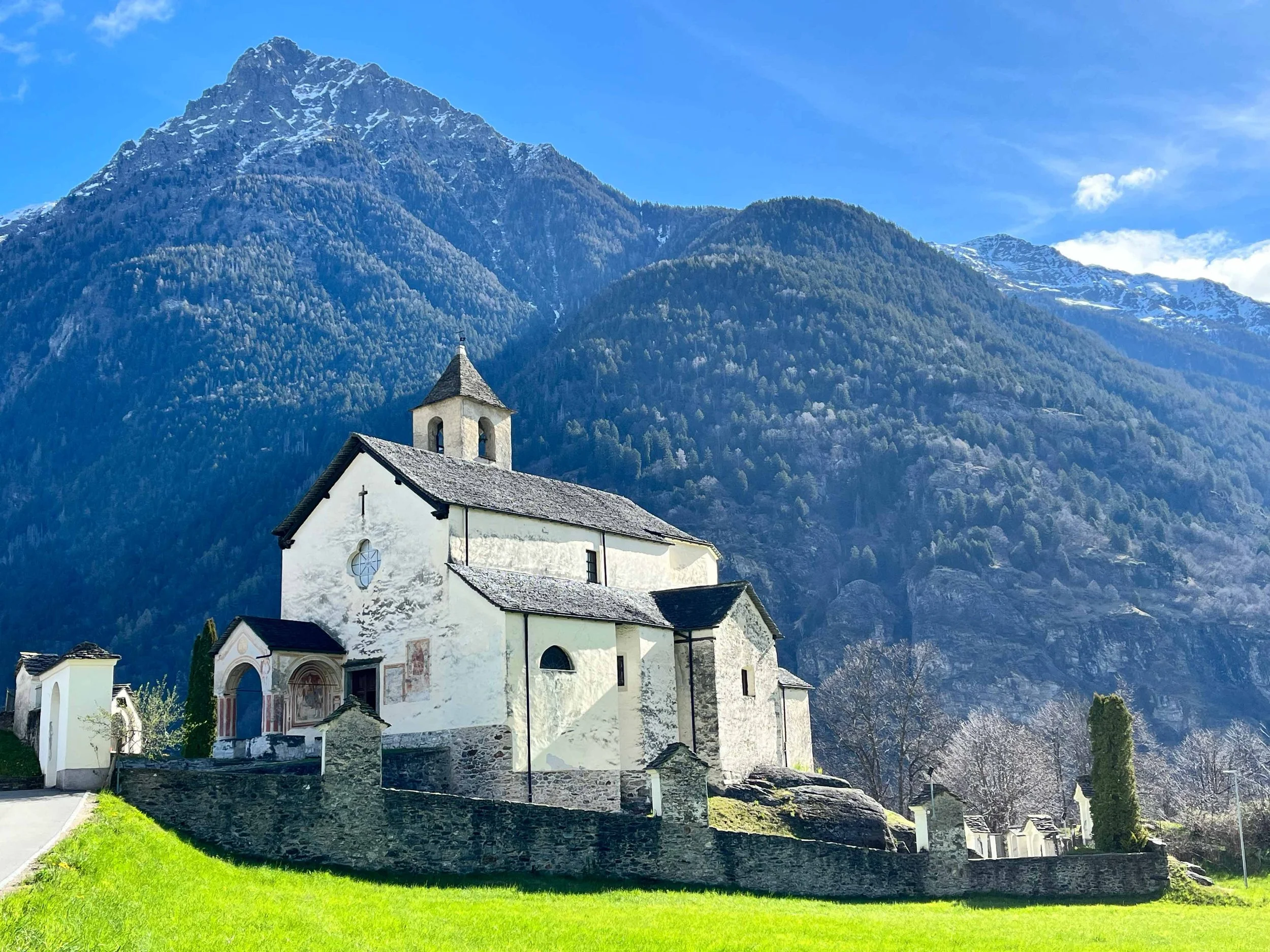 Eine weiße Kirche mit steilem Dach und Glockenturm vor einer Bergkulisse mit hohen, schneebedeckten Gipfeln.