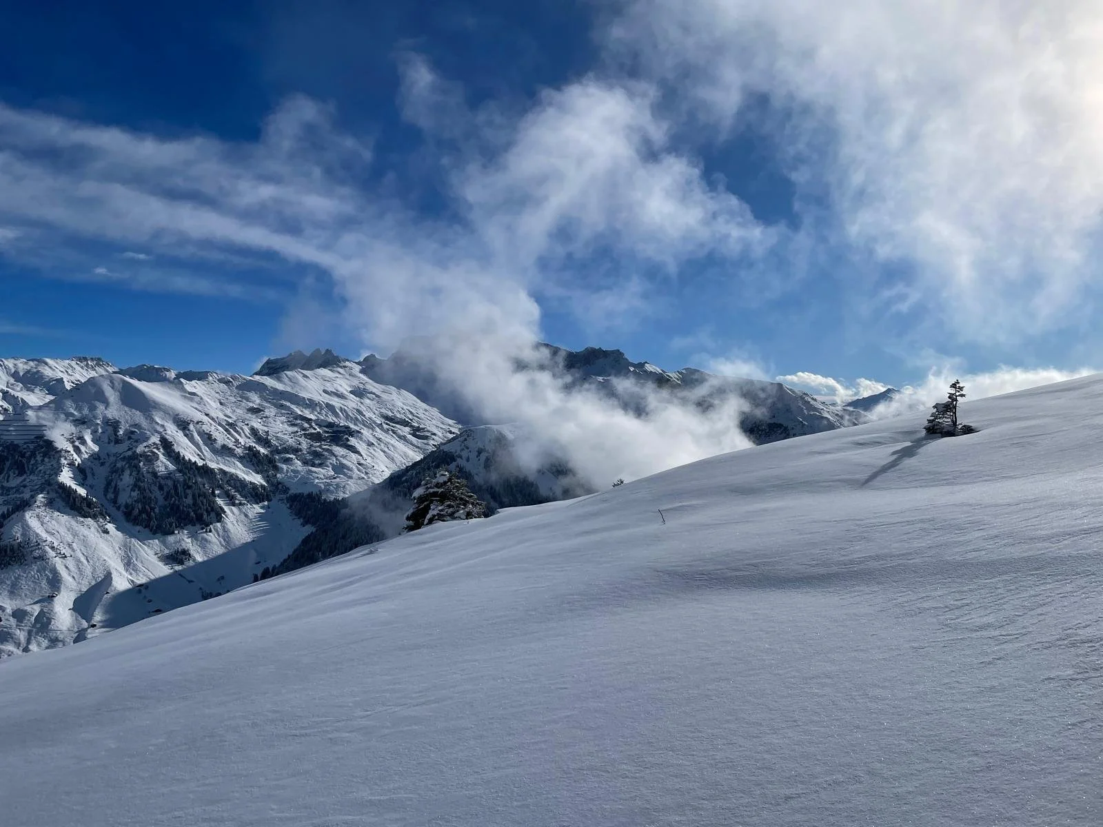 Schneebedeilte Berge unter blauem Himmel mit Wolken und einer einsamen Baumgruppe auf einer weißen Schneefläche im Vordergrund.