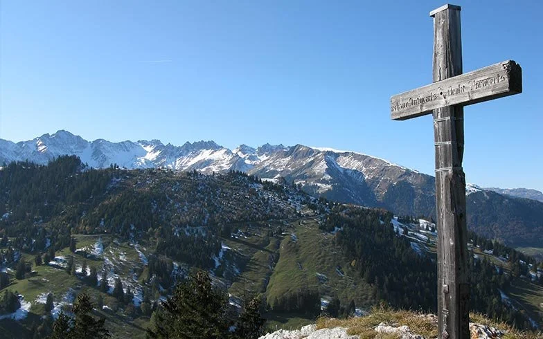 Berglandschaft mit schneebedeckten Gipfeln, bewaldeten Hügeln und einer holzernen Kreuz auf einem Hügel