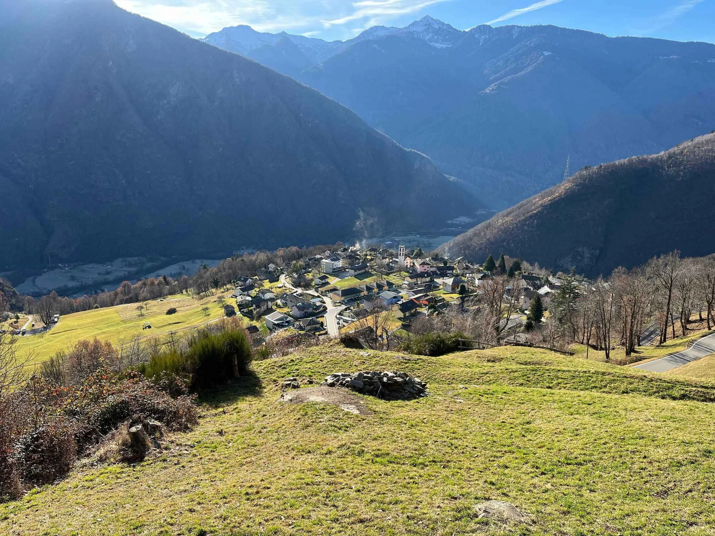 Hügelige Landschaft mit einem kleinen Dorf, umgeben von Bergen im Hintergrund und bewölktem Himmel darüber.