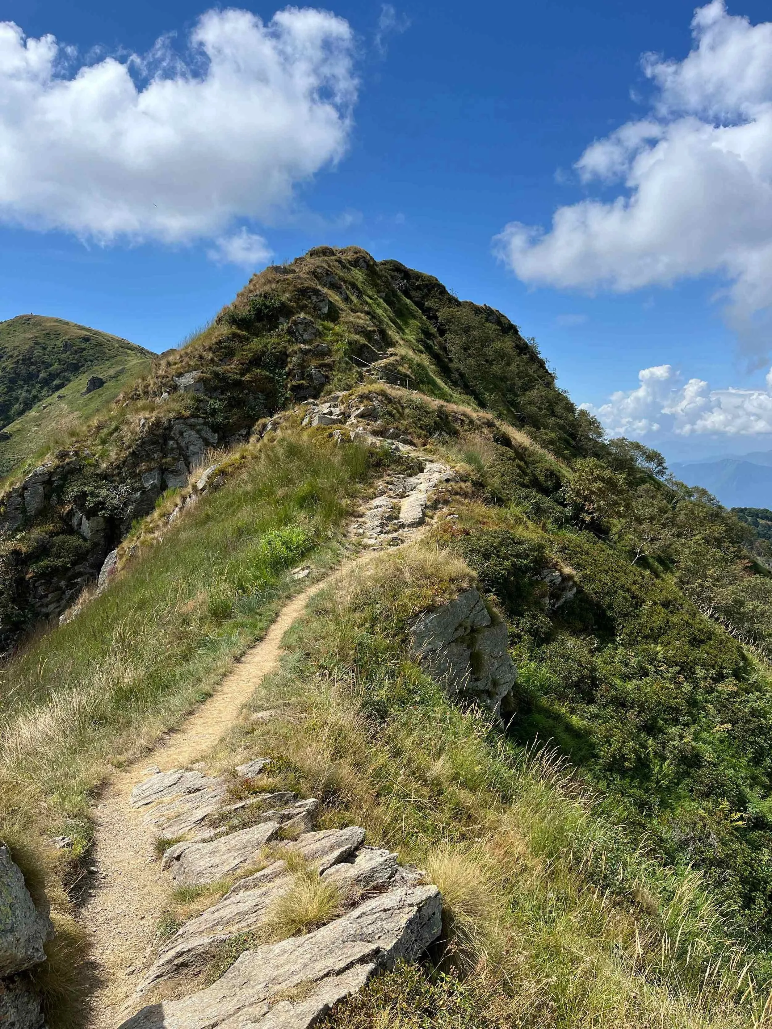 Wanderweg auf einem Bergkamm mit Steinen und Gras, Blick auf den blauen Himmel mit Wolken, in der Ferne Berge
