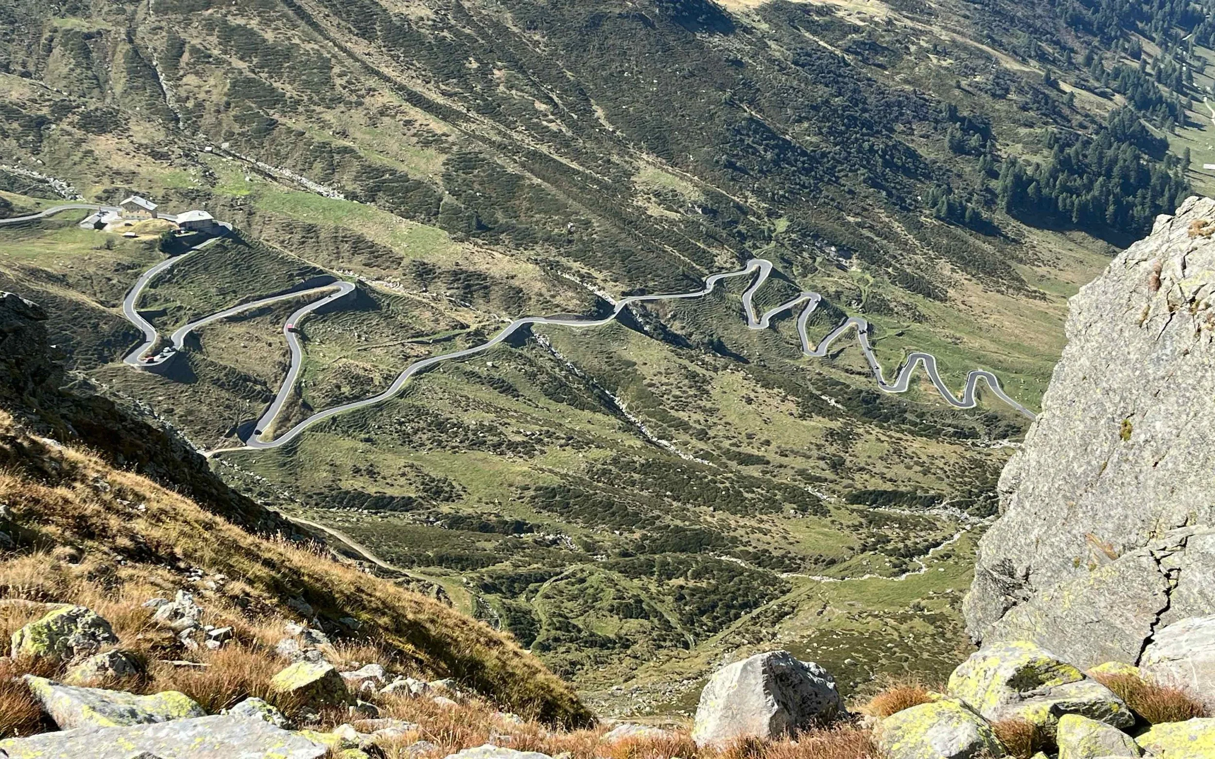 Blick von einer Bergspitze auf eine kurvige Straße in einem grünen, bergigen Gebiet mit wenigen Gebäuden.