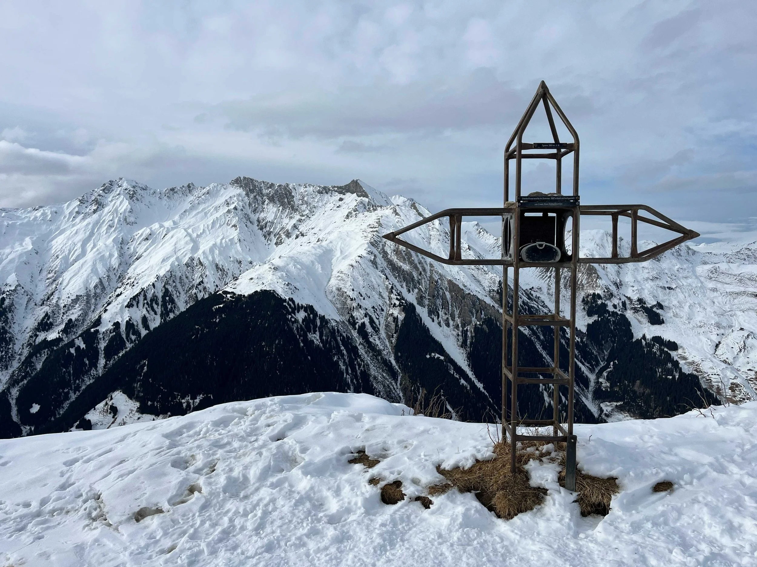 Ein metalldurchbrochener Gipfelkreuz auf schneebedehnten Bergen mit bewölktem Himmel im Hintergrund.