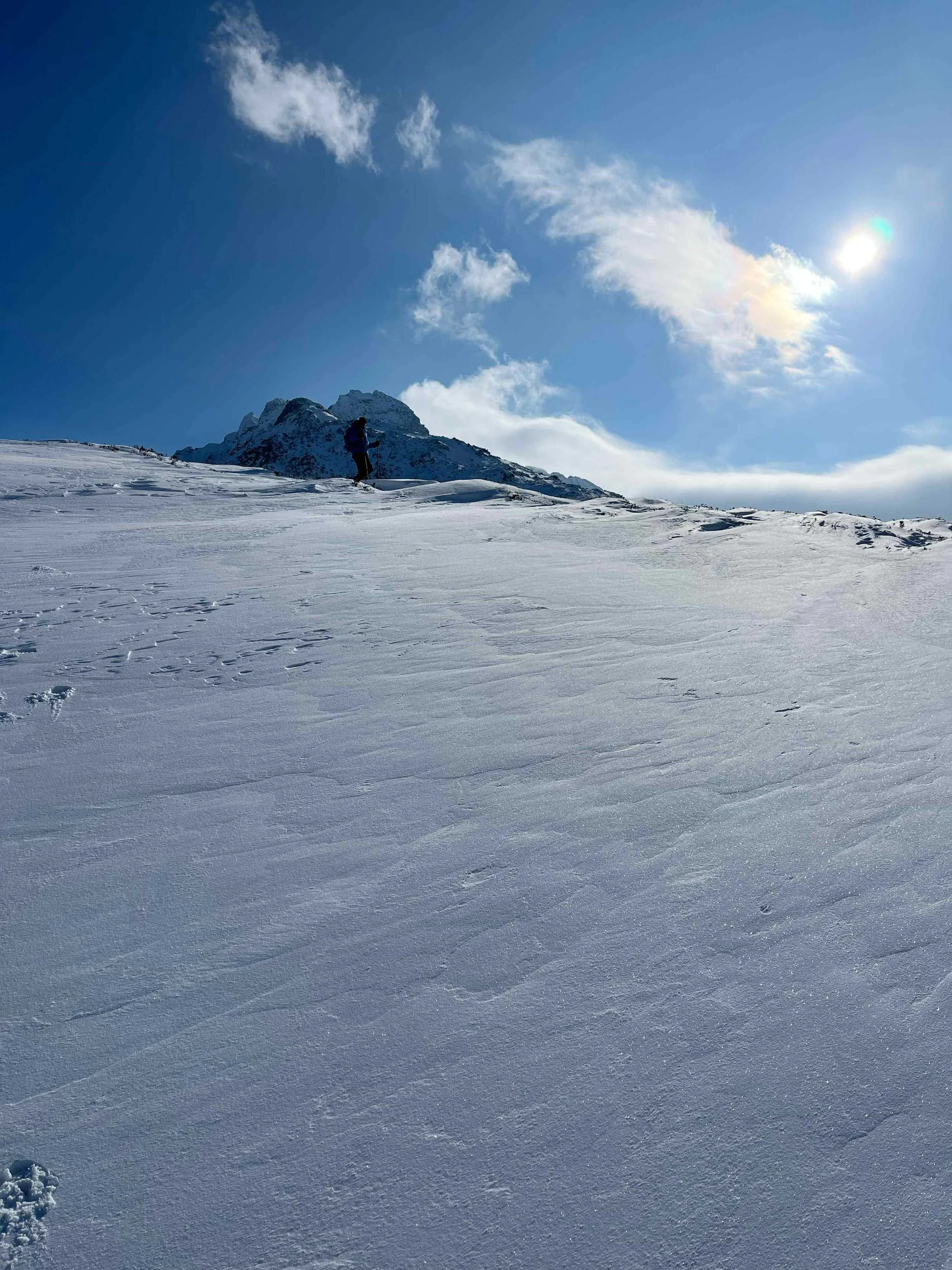 Schneebedecktes Bergpanorama mit einem einzelnen Skifahrer an der Spitze und einem klaren blauen Himmel mit wenigen Wolken und der Sonne im Hintergrund.