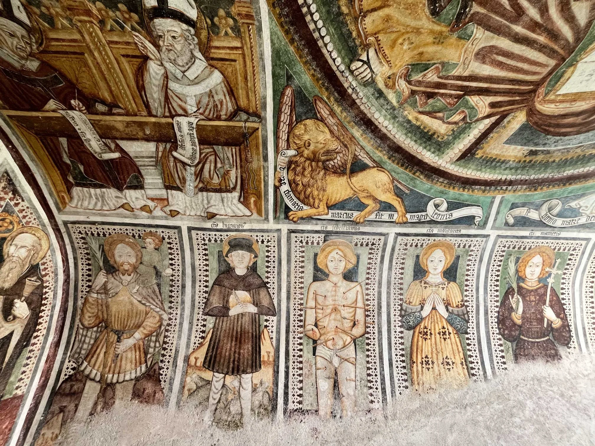 Fresko mit verschiedenen Heiligen und biblischen Szenen, inklusive eines Löwen, in einer Kirche oder Kapelle. Der Stil ist gotisch, mit detaillierten, farbigen Darstellungen.