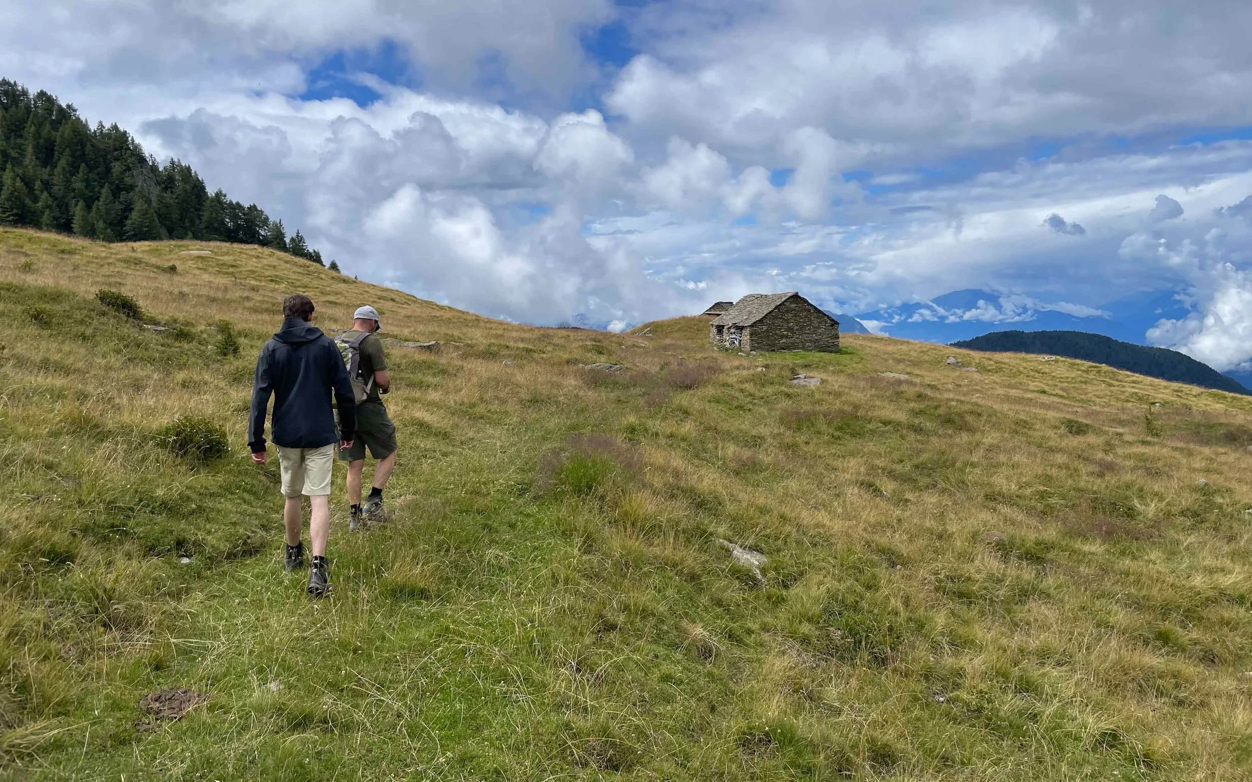 Zwei Wanderer auf einer grünen Bergwiese mit einem kleinen steinernen Haus im Hintergrund und einem bewölkten Himmel.