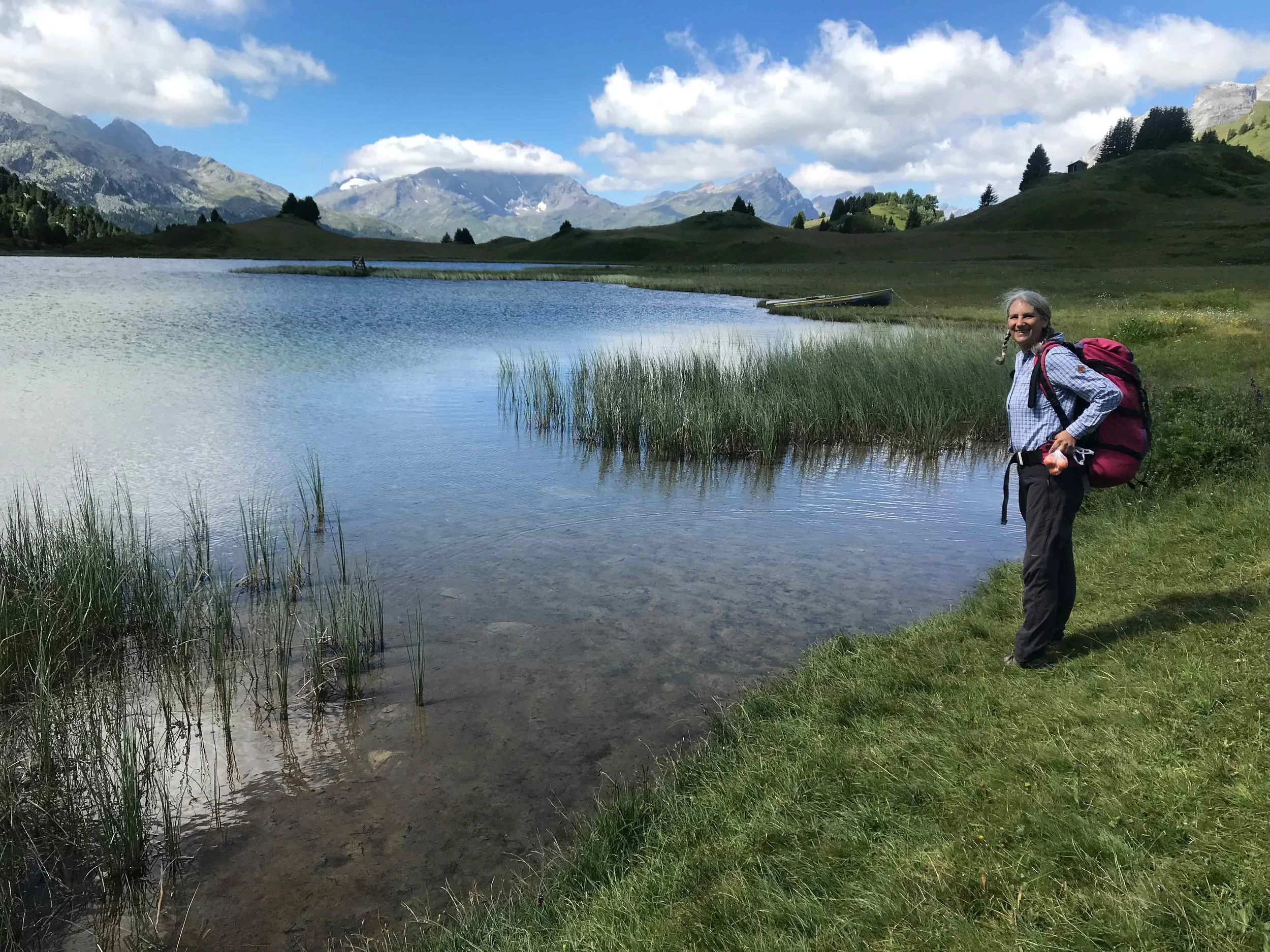 Junge Frau mit Rucksack am See in den Bergen, bewölkter Himmel, grüne Wiesen und Berge im Hintergrund.