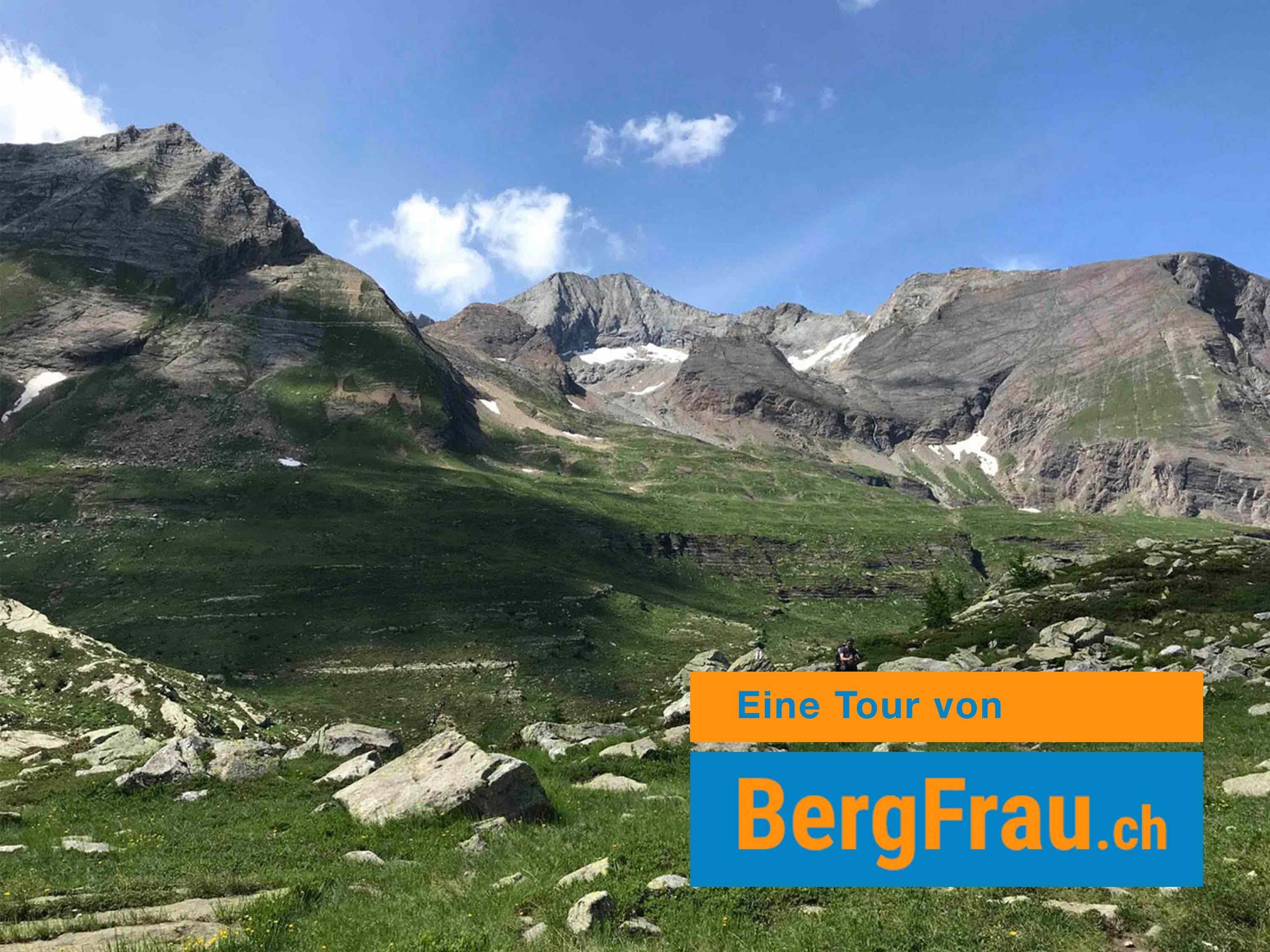 Berglandschaft mit hohen Gipfeln, grünen Wiesen und einzelnen Wolken am blauen Himmel.