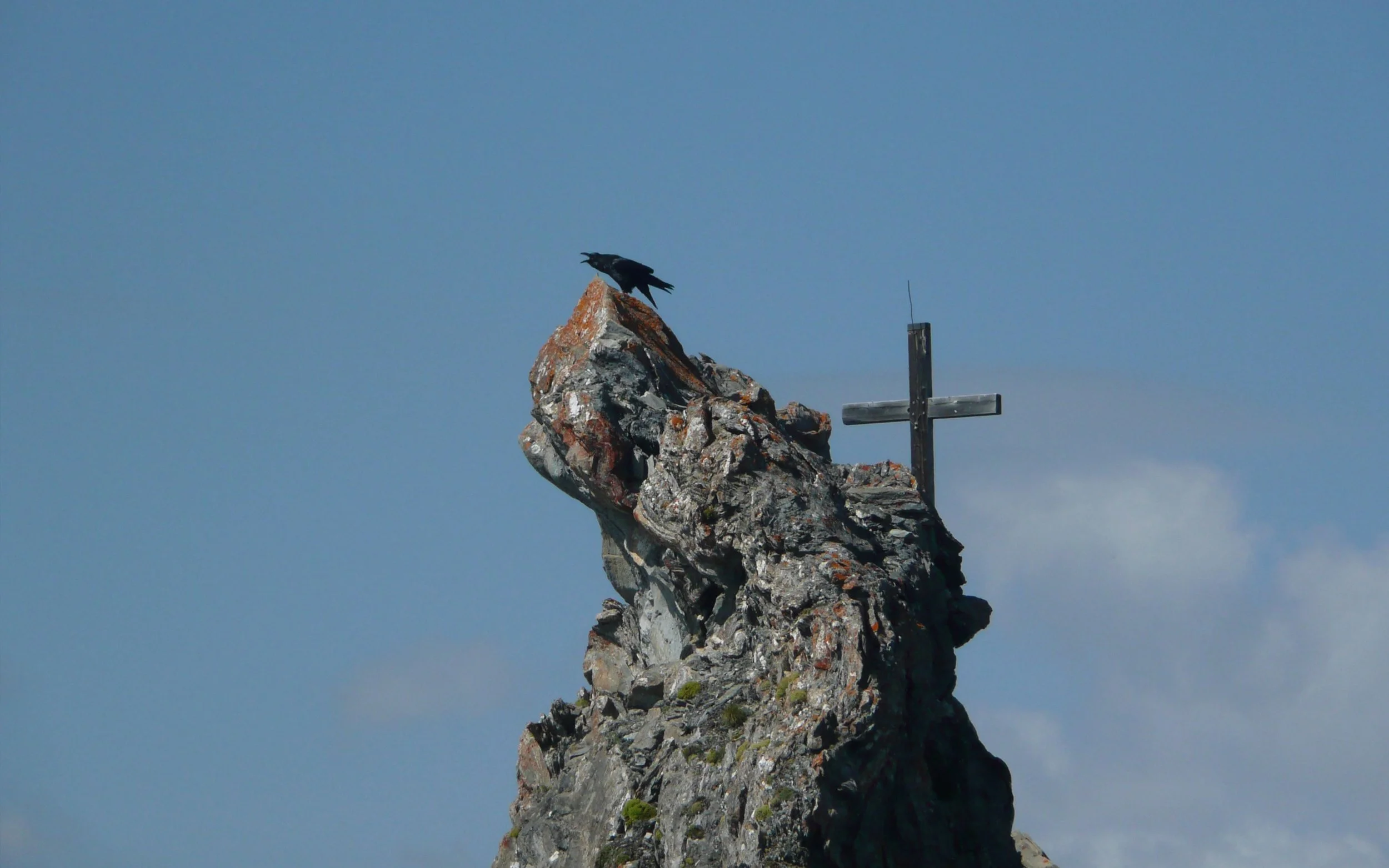 Ein dunkler Vogel auf einem felsigen Gipfel mit einem Holzkreuz im Hintergrund, Himmel mit wenigen Wolken.