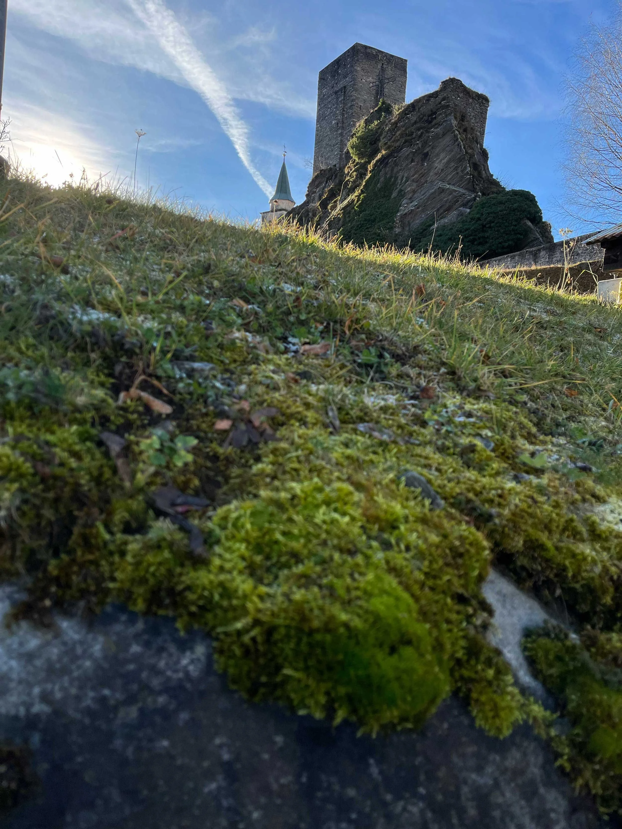 Ein Blick aus niedriger Perspektive auf die Grasfläche im Vordergrund, mit einer alten Burg oder Festung auf einem Felsen im Hintergrund, bei blauem Himmel und einigen Wolken.