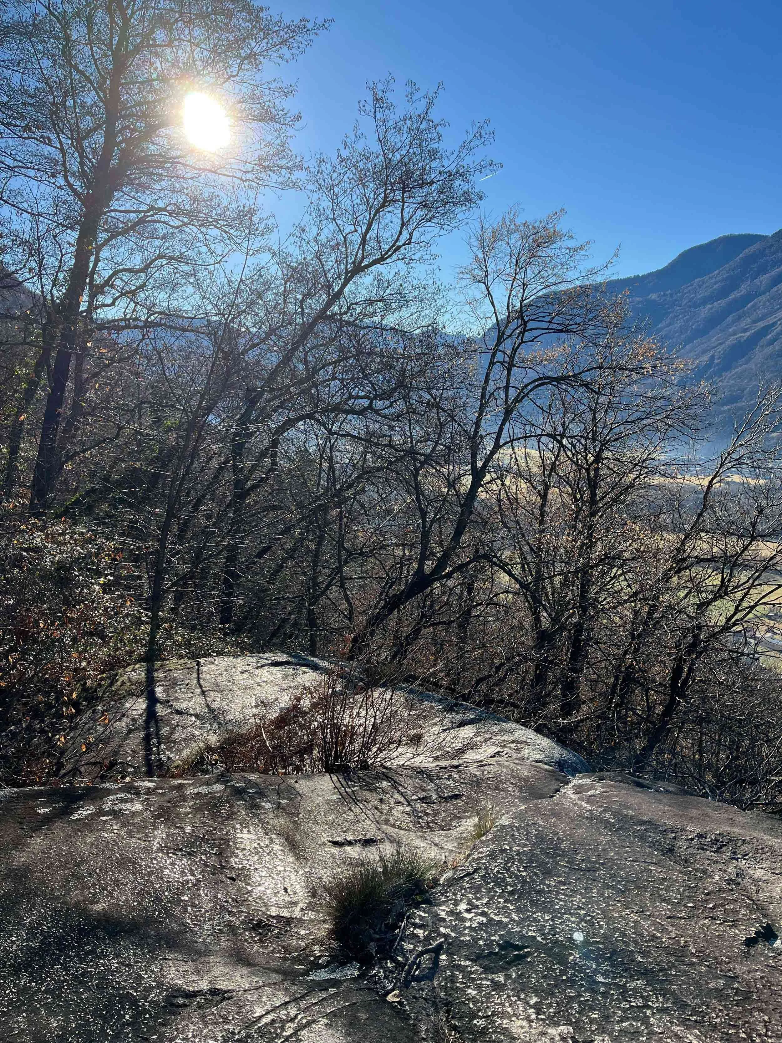 Berglandschaft mit Bäumen, Sonne, Himmel und Wasser im Vordergrund.