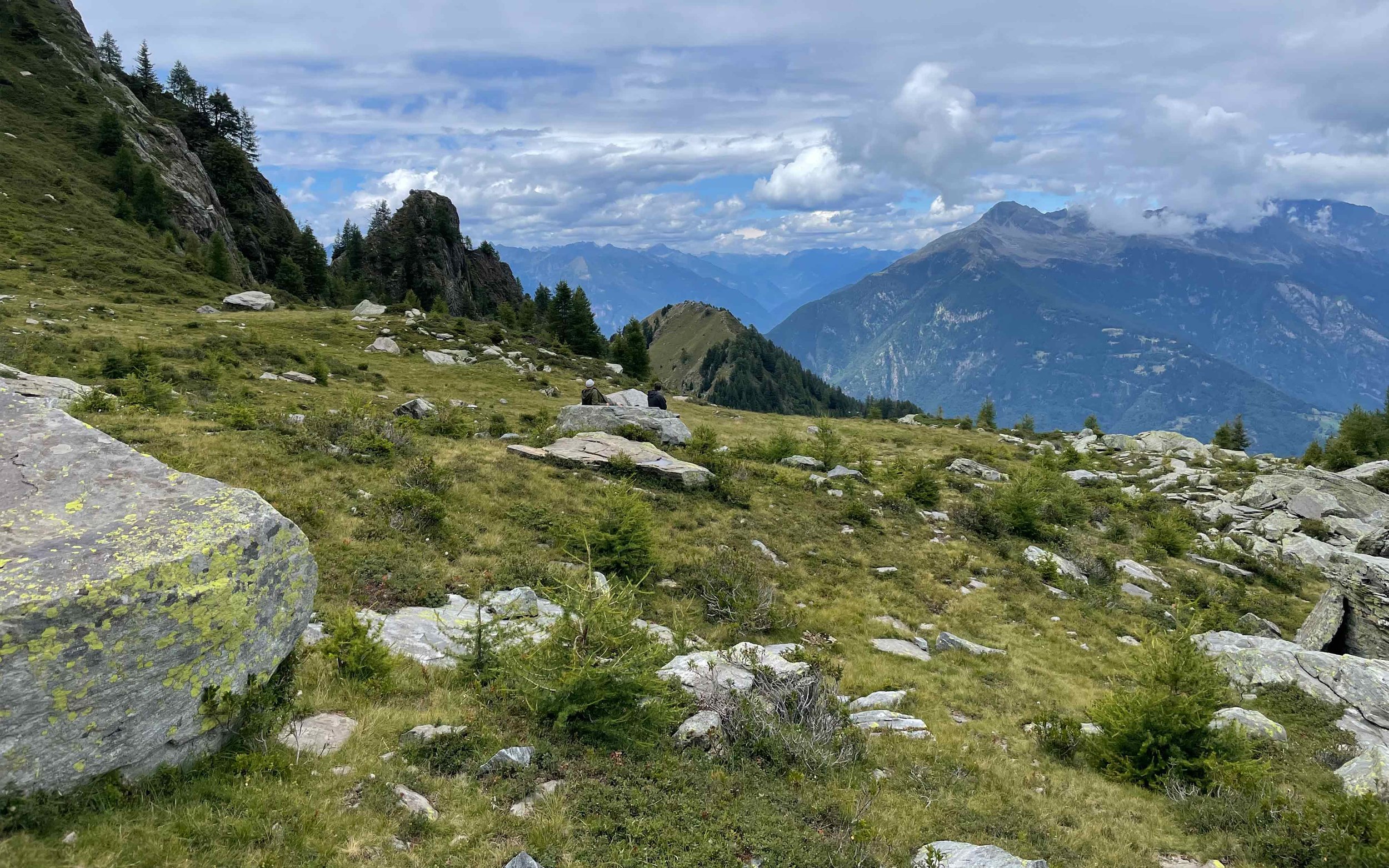 Berglandschaft mit grünen Wiesen, Felsen und2 bis 3 Wanderern, im Hintergrund hohe Berge unter bewölktem Himmel.