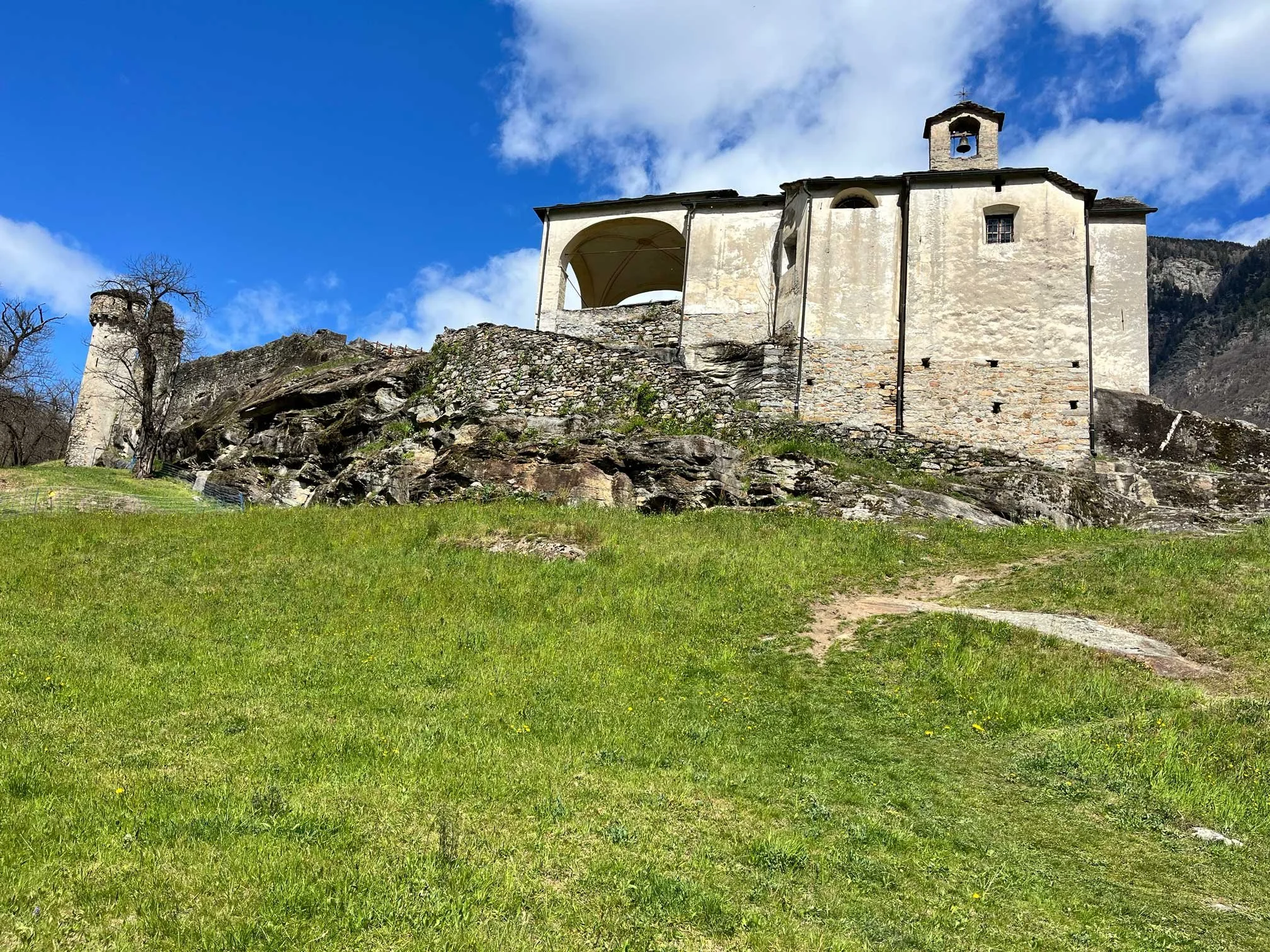 Alte Burg auf einem Felsen in einer grünen Wiese, im Hintergrund Himmel mit Wolken.