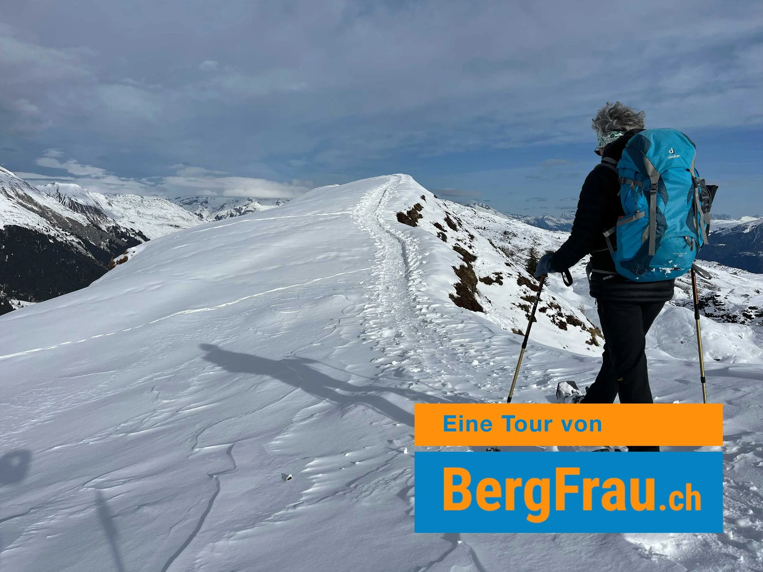 Eine Person mit Skiern, die auf einem schneebedeckten Berggipfel steht, trägt eine blaue Jacke, eine Skibrille, eine Kopfbedeckung und einen Rucksack. Der Himmel ist bewölkt mit blauen und grauen Wolken.
